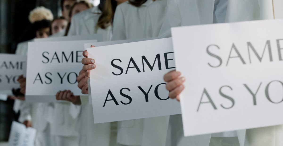 Several people in white suits hold up signs that say, "SAME AS YOU."