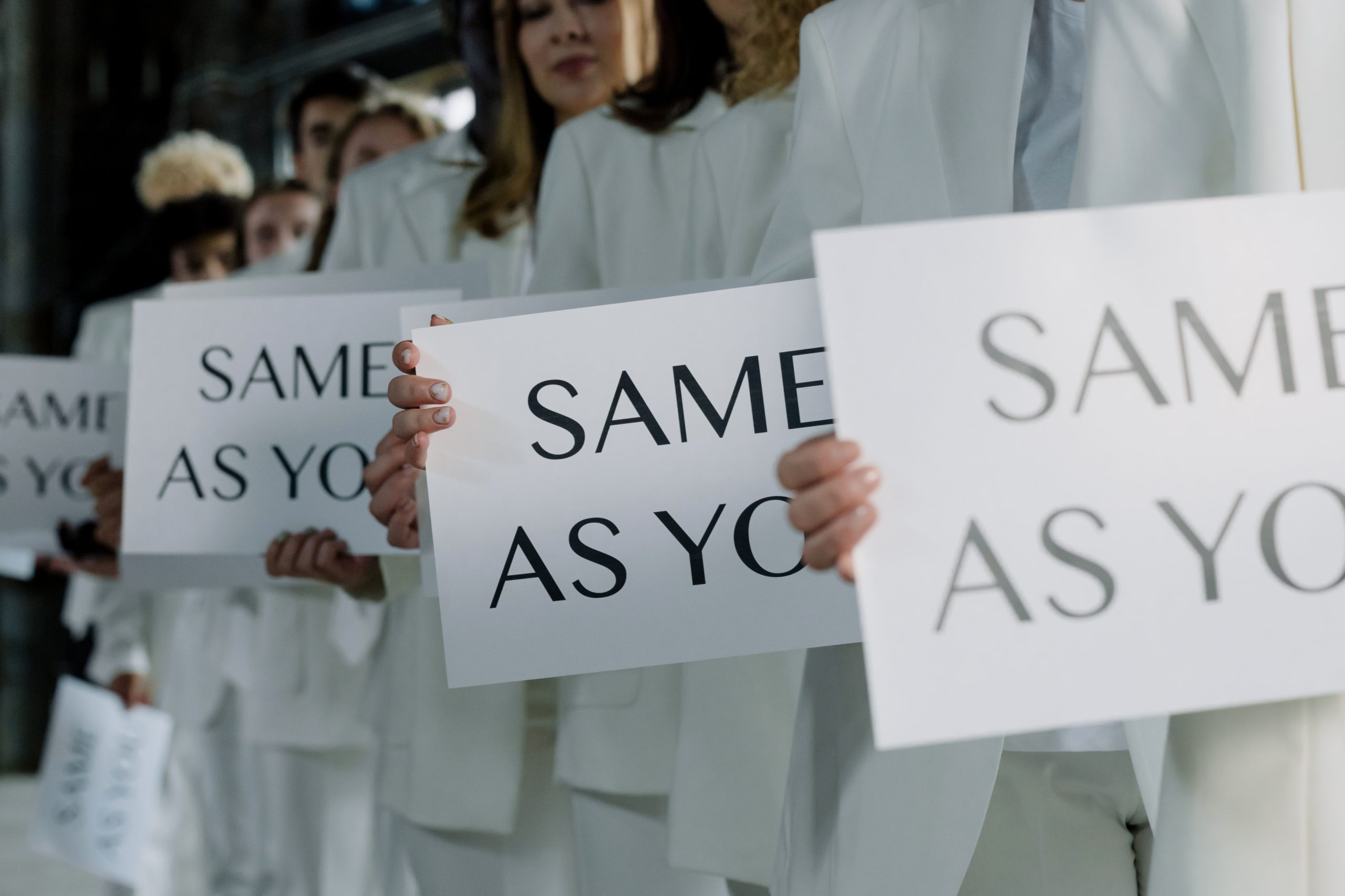 Several people in white suits hold up signs that say, "SAME AS YOU."