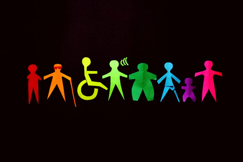 Some paper cut-outs of various people on different colours lie on a black surface. The people include a person with missing arm limbs, a visually impaired person, a person in a wheelchair, an obese person, a person with a prosthetic leg and a dwarf.