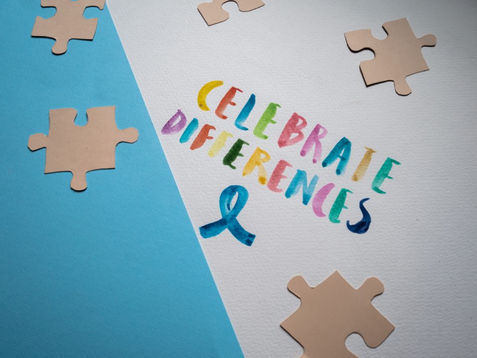 The slogan, "CELEBRATE DIFFERENCES," with each letter a different colour and a blue ribbon are painted on a white sheet of paper. The white sheet is lying on top of a blue sheet of paper. Some brown paper puzzle pieces lie on both sheets of paper.