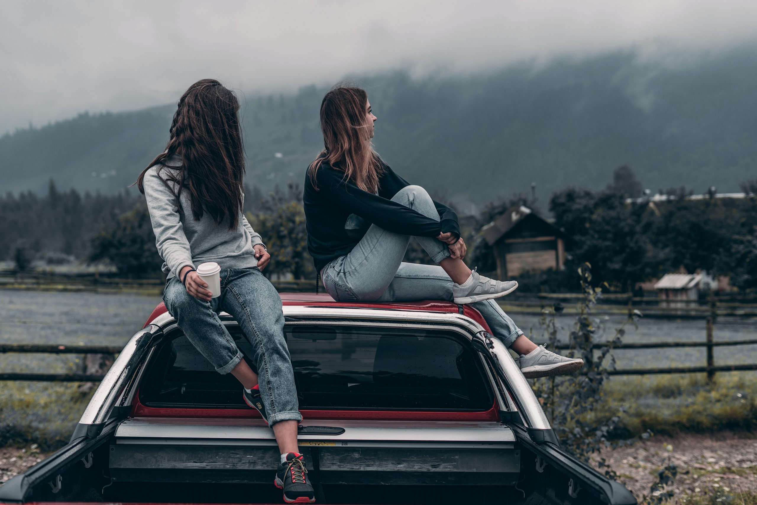 Two women sit on top of a car. One woman looks at the other, but the other woman looks away.