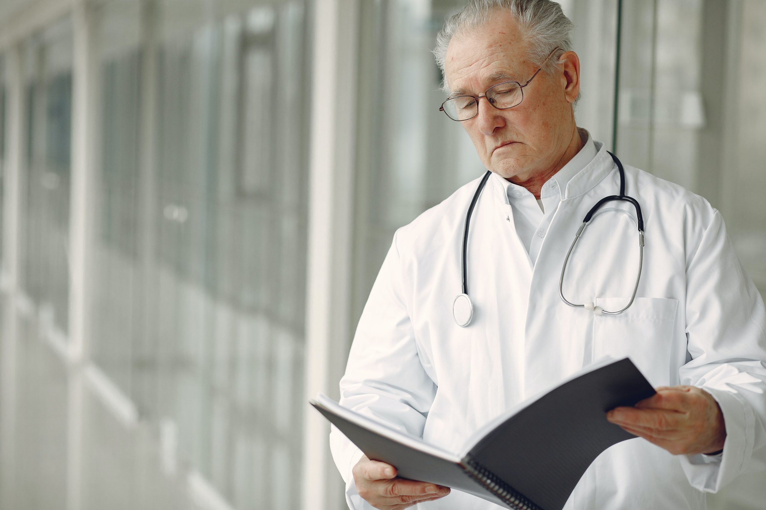A middle-aged to elderly male doctor is holding an open book and looking down on it.