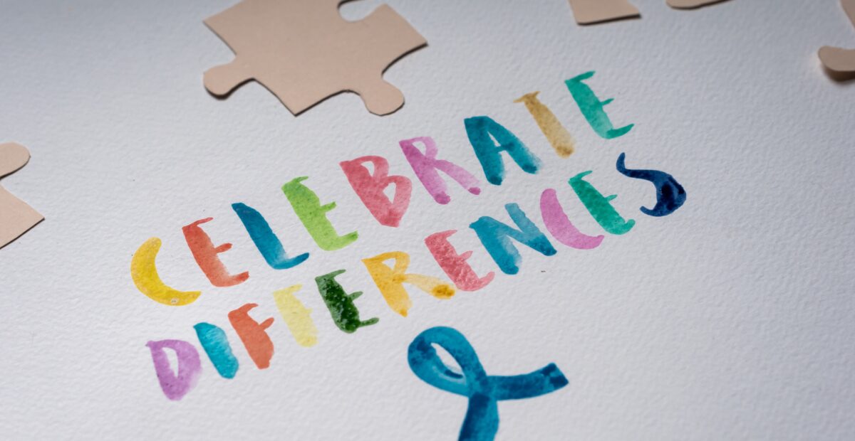 The words, "CELEBRATE DIFFERENCES," in various colours and a blue ribbon are painted on some white paper. Some brown paper puzzle pieces lie on the white paper.