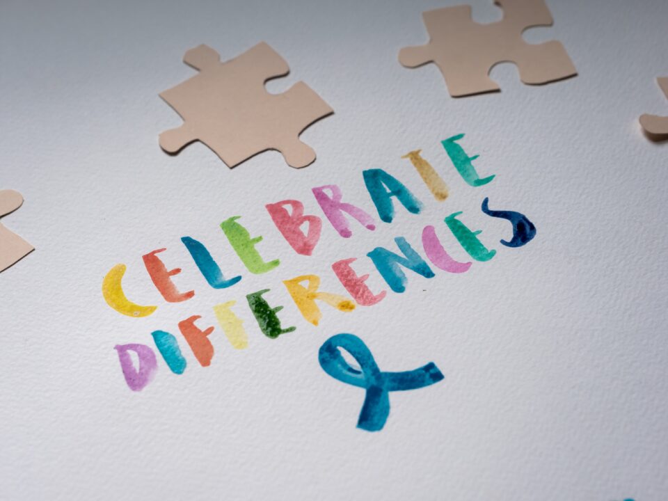 The words, "CELEBRATE DIFFERENCES," in various colours and a blue ribbon are painted on some white paper. Some brown paper puzzle pieces lie on the white paper.
