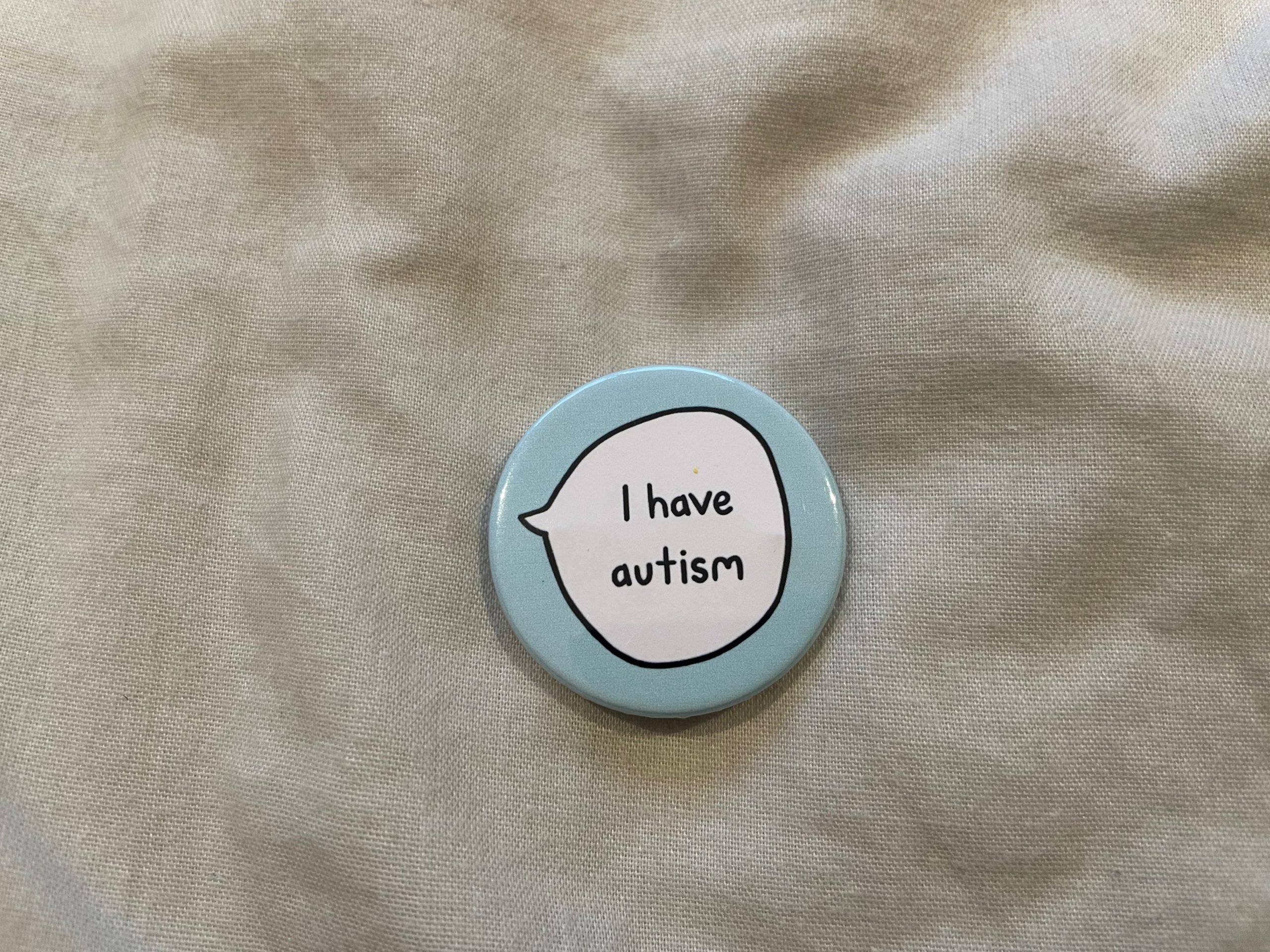 A pale blue badge with a speech bubble that says, "I have autism,' lies on a light grey bedsheet.