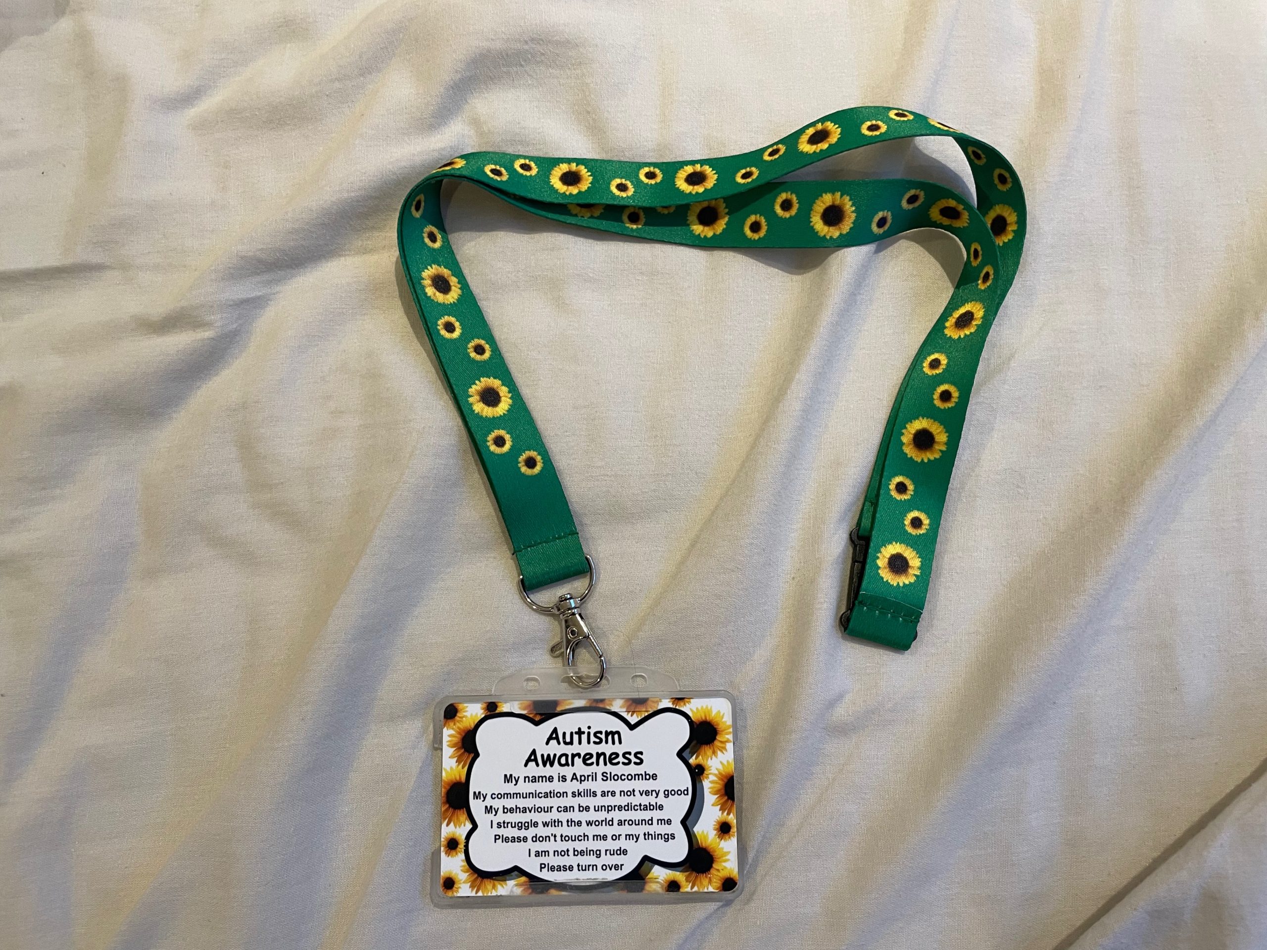 An autism awareness card with a sunflower border is attached to a green lanyard with sunflowers on it. The card and the lanyard are lying on a light grey bedsheet.