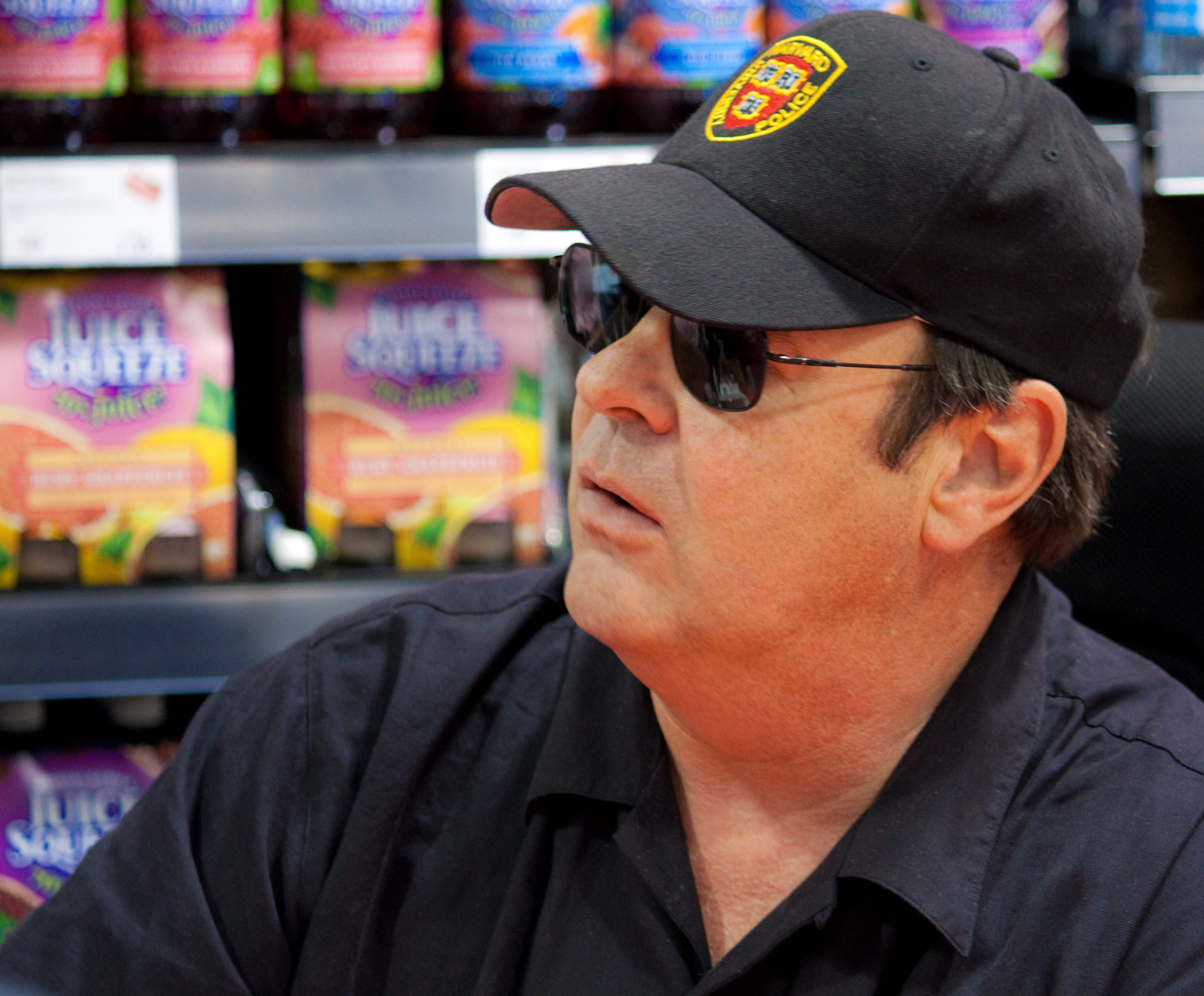 The actor Dan Aykroyd is sitting in a record shop. He is wearing a baseball cap, some sunglasses and a dark polo shirt.