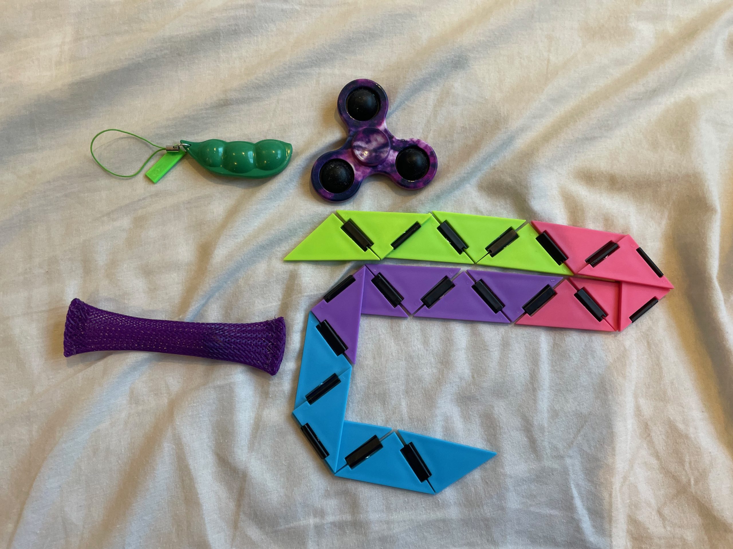 Four fidget toys lie on a grey bedsheet. They are green peas in a pod, a purple fidget spinner, a snake-like multicoloured toy and a purple mesh tube.