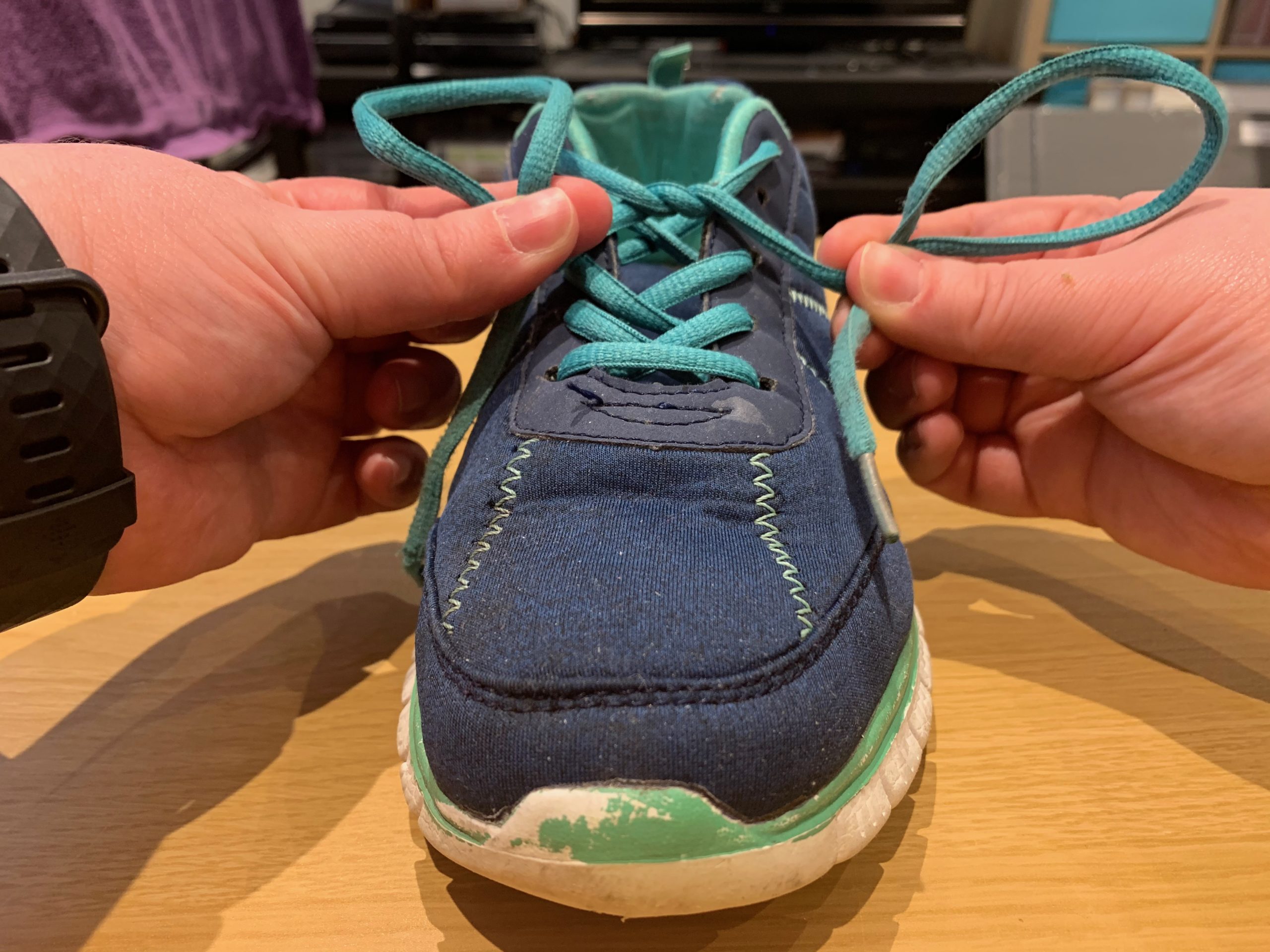 A navy and teal trainer with hands holding its laces into loops.