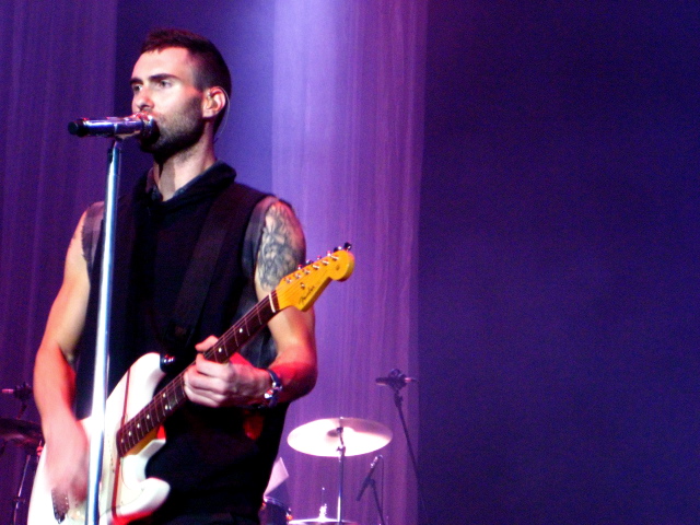 Adam Levine, lead singer of Maroon 5, is singing and playing the guitar in front of a stripy purple background.