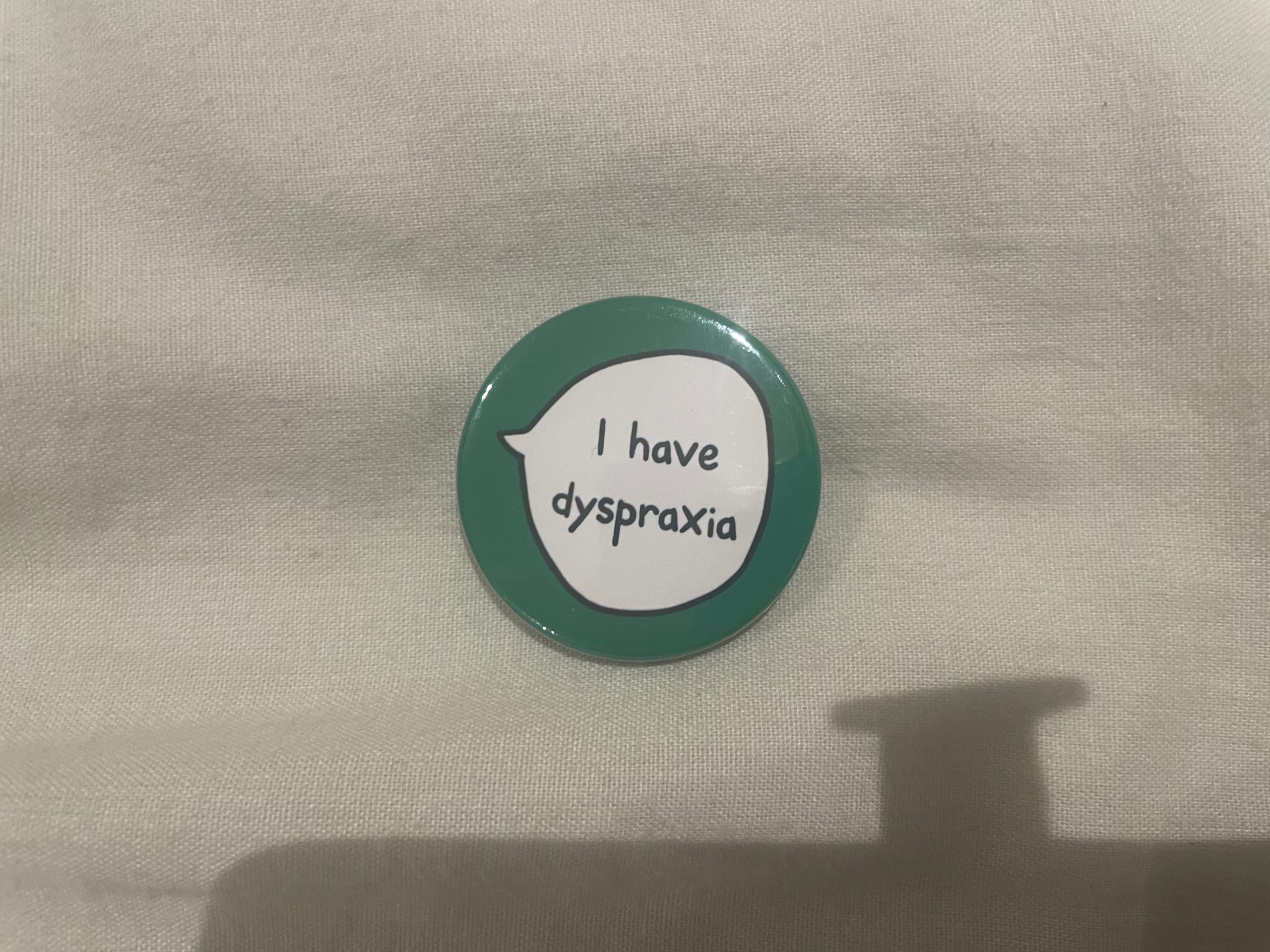 A green badge with a speech bubble that says, "I have dyspraxia," lies on a grey bedsheet.