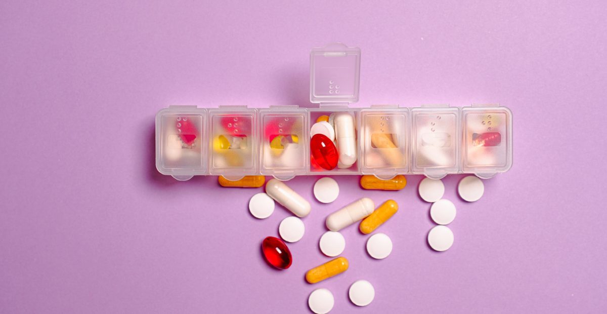 A plastic container with multiple sections, some tablets and capsules lie on a lilac surface.