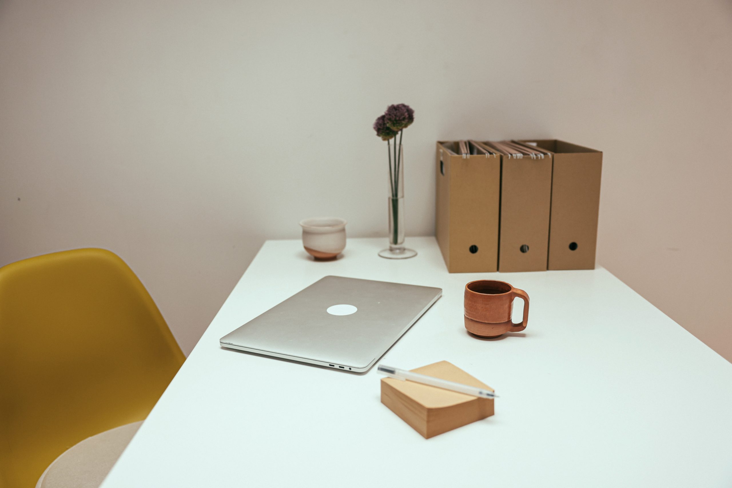 A white desk has some magazine file boxes, a glass vas of fake flowers, a small ceramic vase, a laptop, a mug, some sticky notes and a pen lying on it. There is a yellow plastic chair under the desk.