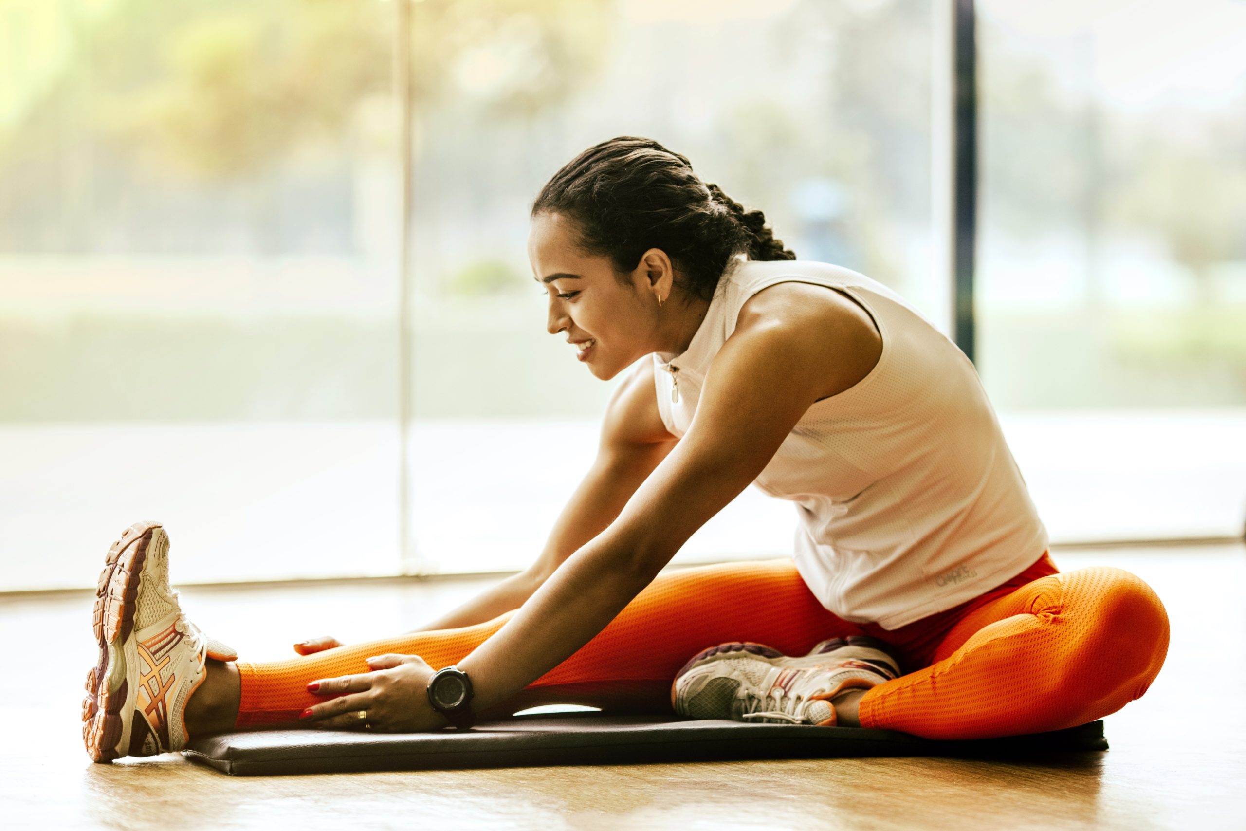 A woman is sitting on an exercise mat. She is stretching one leg out straight and she has the other one bent towards her. She is holding her stretched leg.