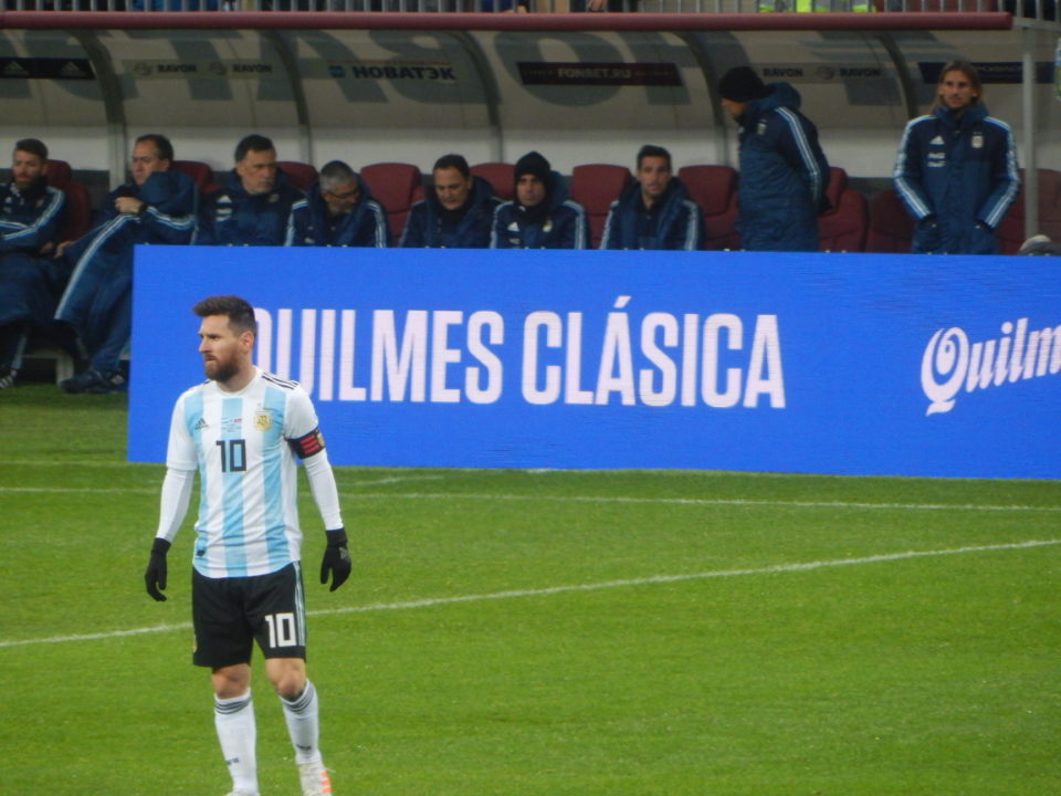 Lionel Messi is standing on a football pitch. Behind him are a blue banner and some men sitting on a row of chairs.