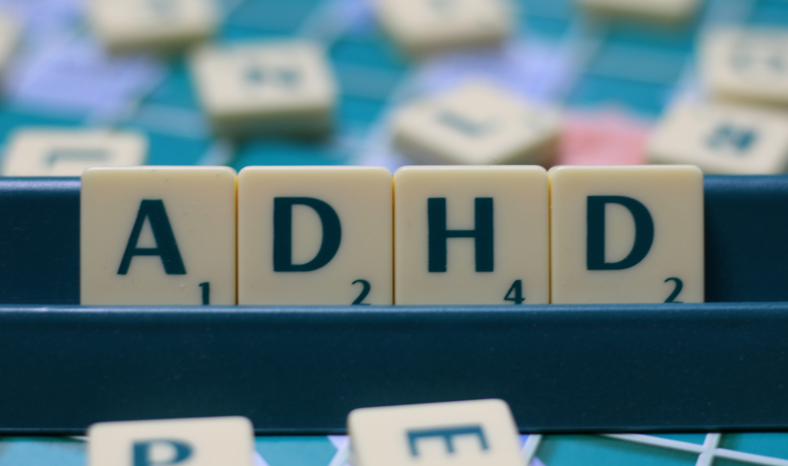 Some Scrabble tiles spell out, "ADHD," on a tile holder. Some more Scrabble tiles and a Scrabble board can be seen in the background.