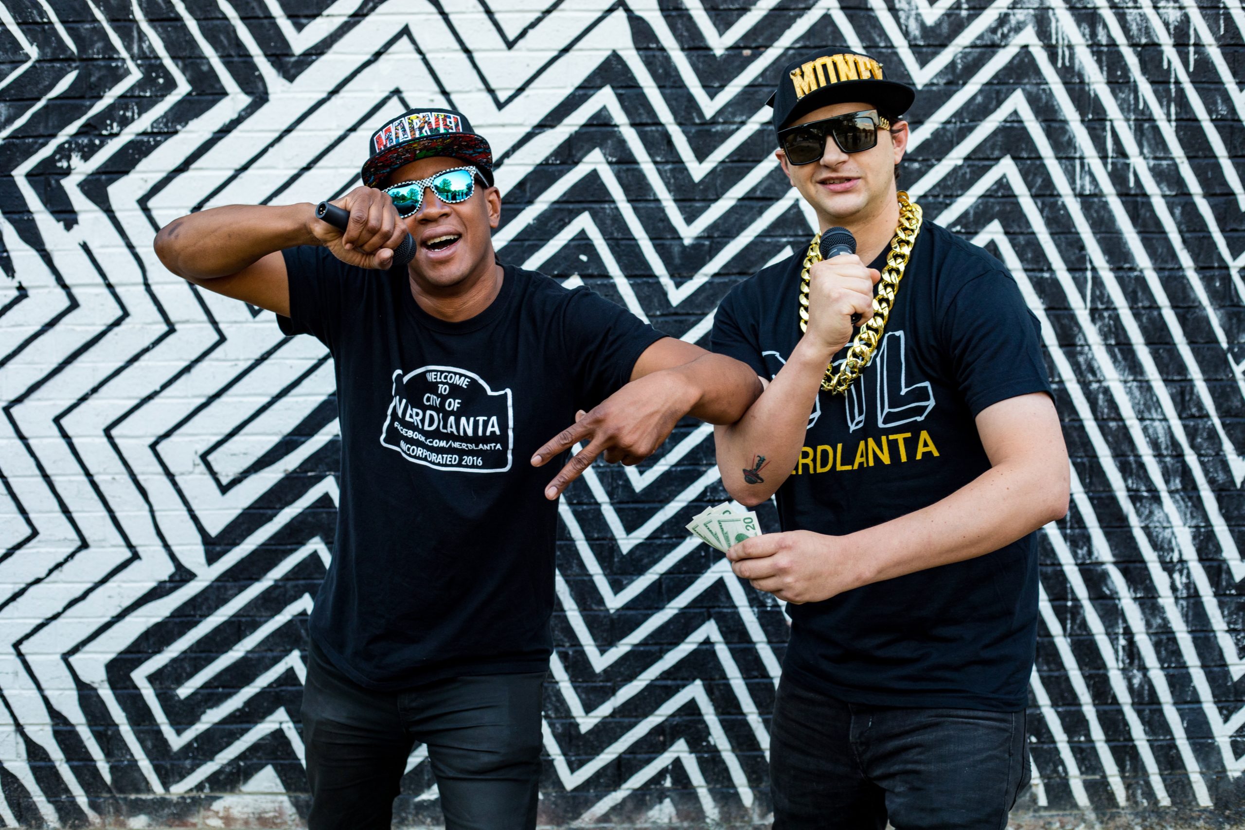 Two rappers perform in front of a brick wall with a black and white zig zag pattern on it. The rapper on the right is holding some banknotes in one hand.