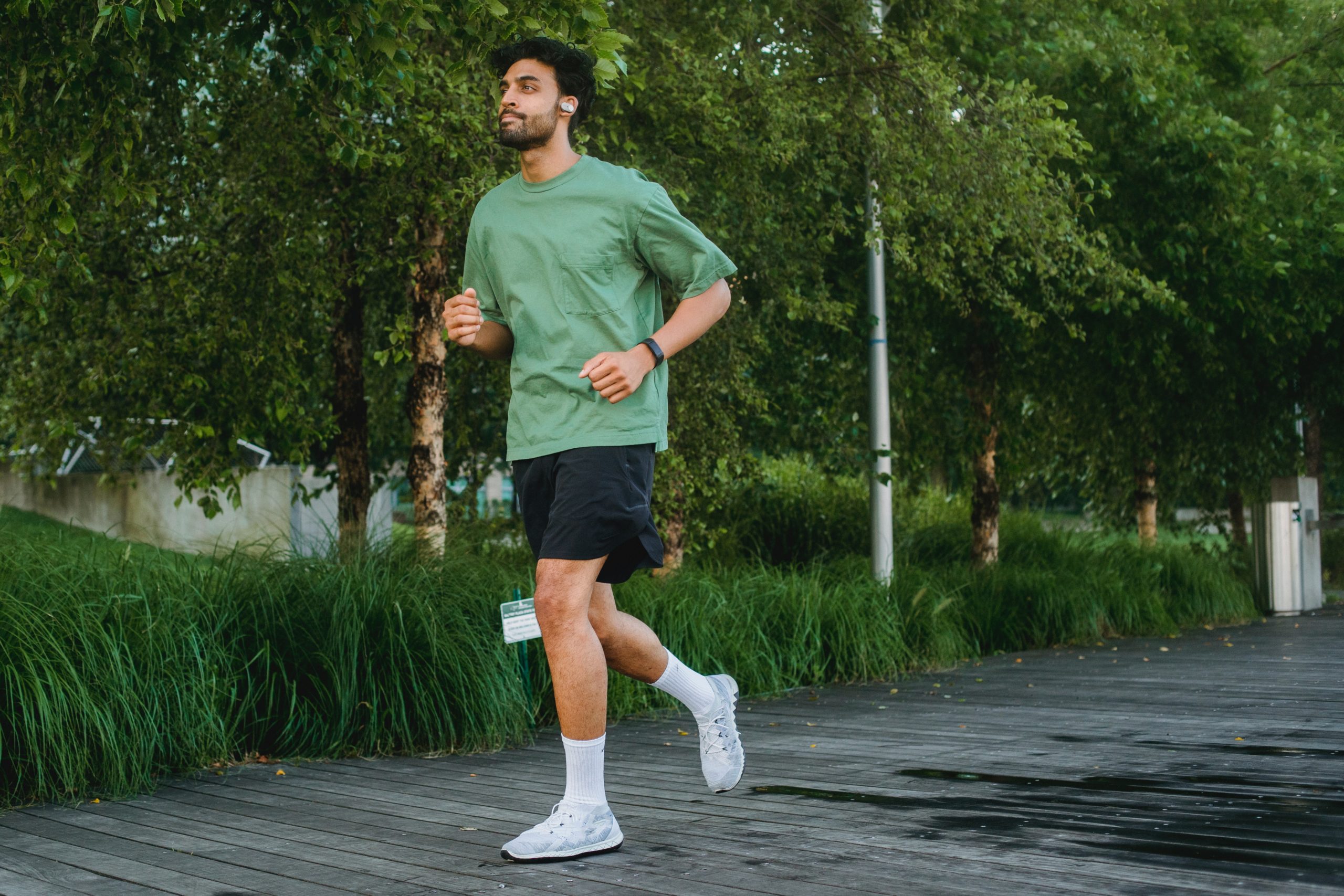 A man in a green T-shirt, black shorts, white socks and white trainers is running past some trees.