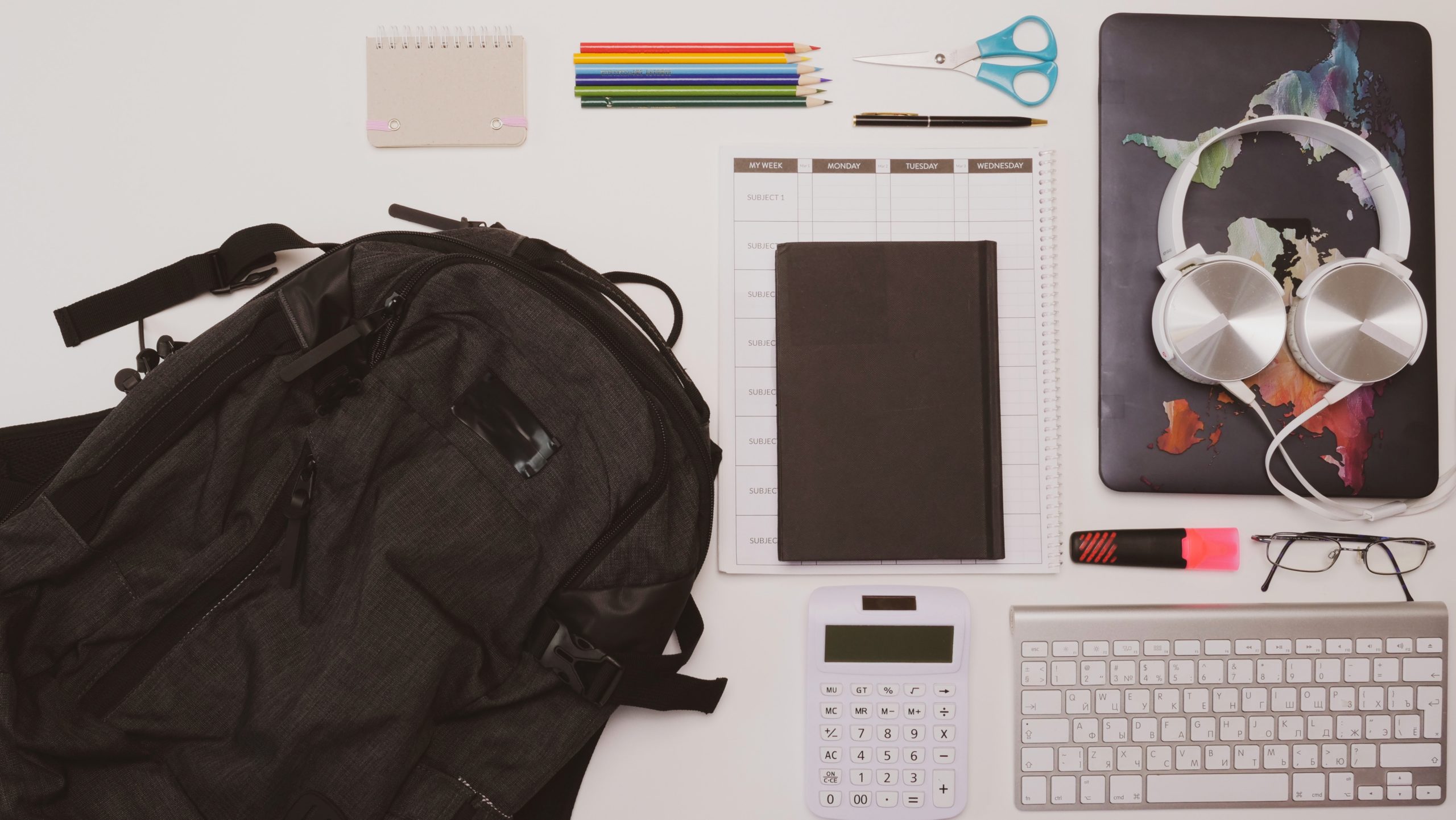 A backpack and several pieces of school equipment, a laptop, some headphones and a computer keyboard lie on a white surface.