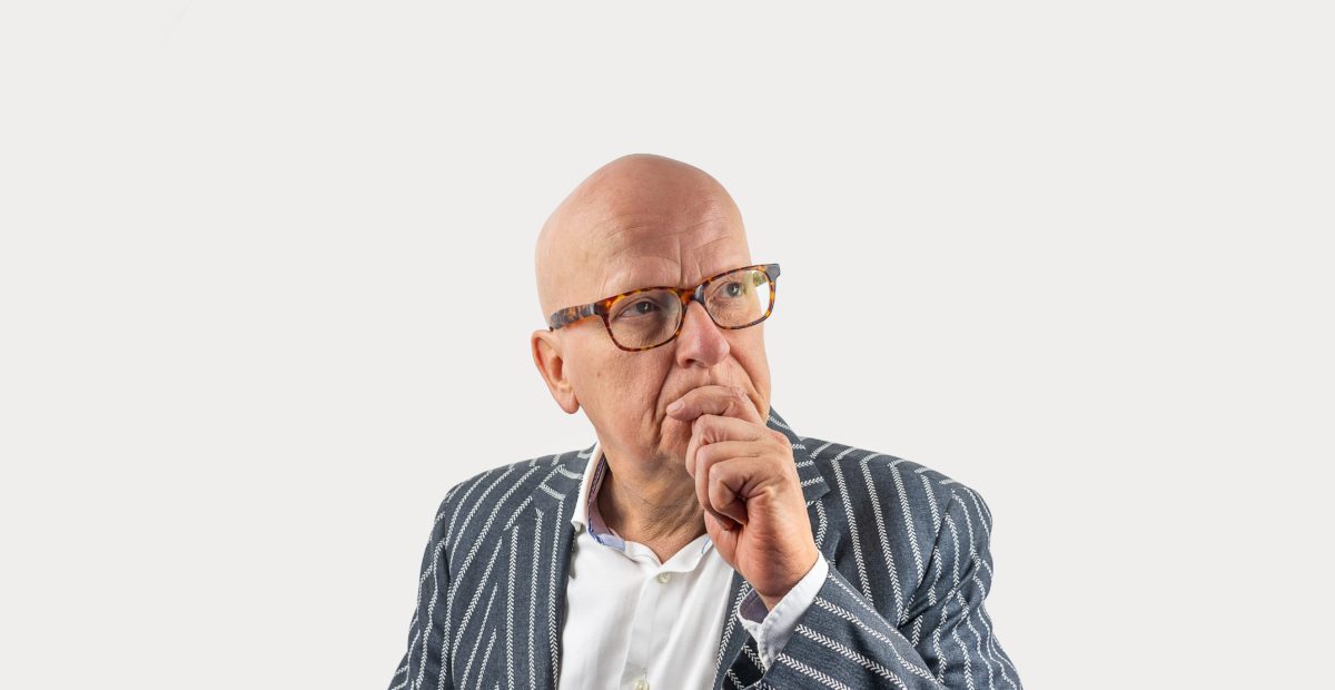 A bald elderly man in glasses and a stripy suit looks deep in thought.