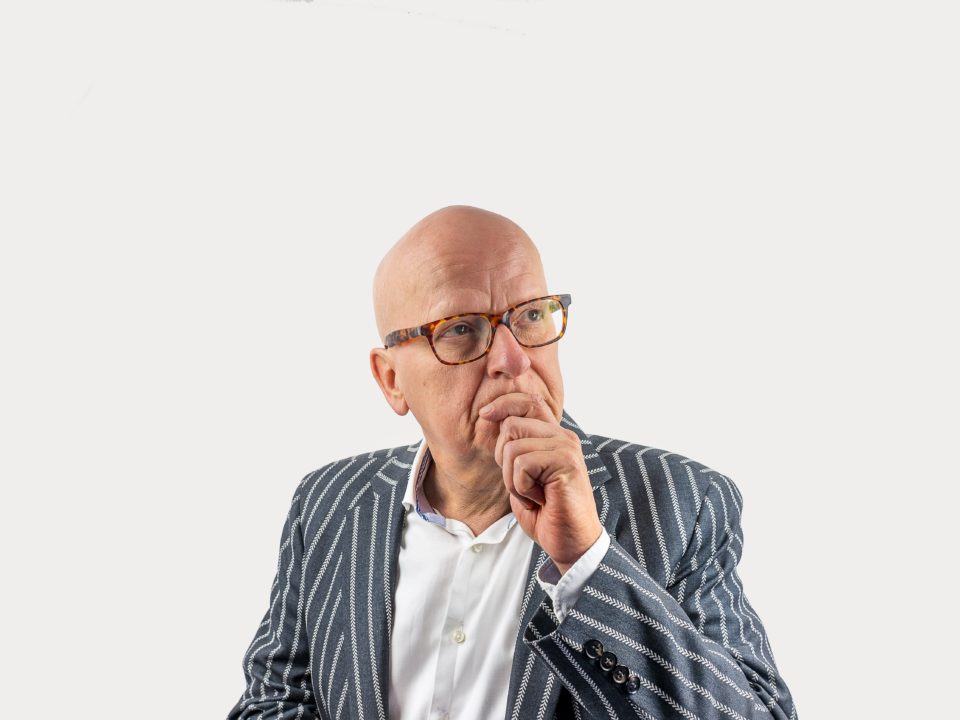 A bald elderly man in glasses and a stripy suit looks deep in thought.