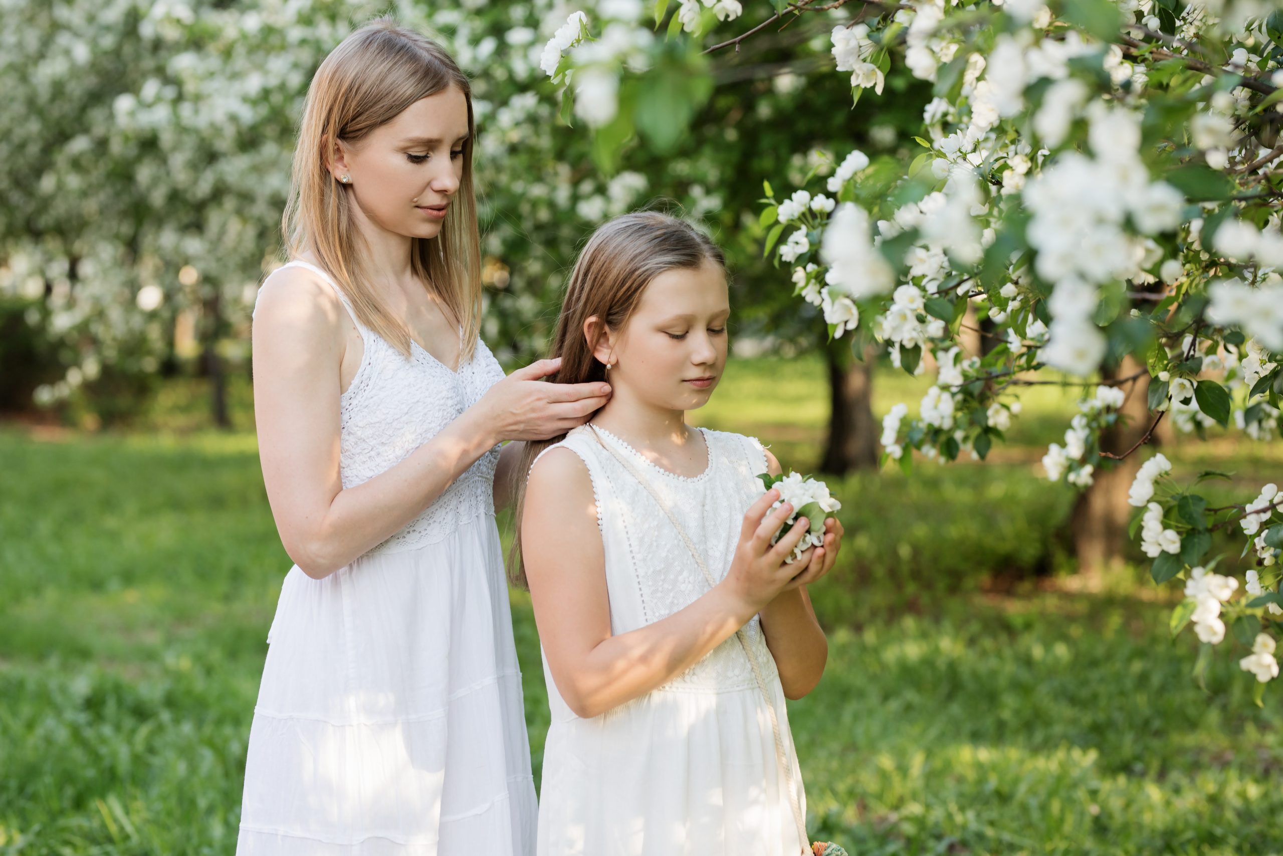 A mother and a daughter are wearing white dresses. The mother is styling the daughter's hair.