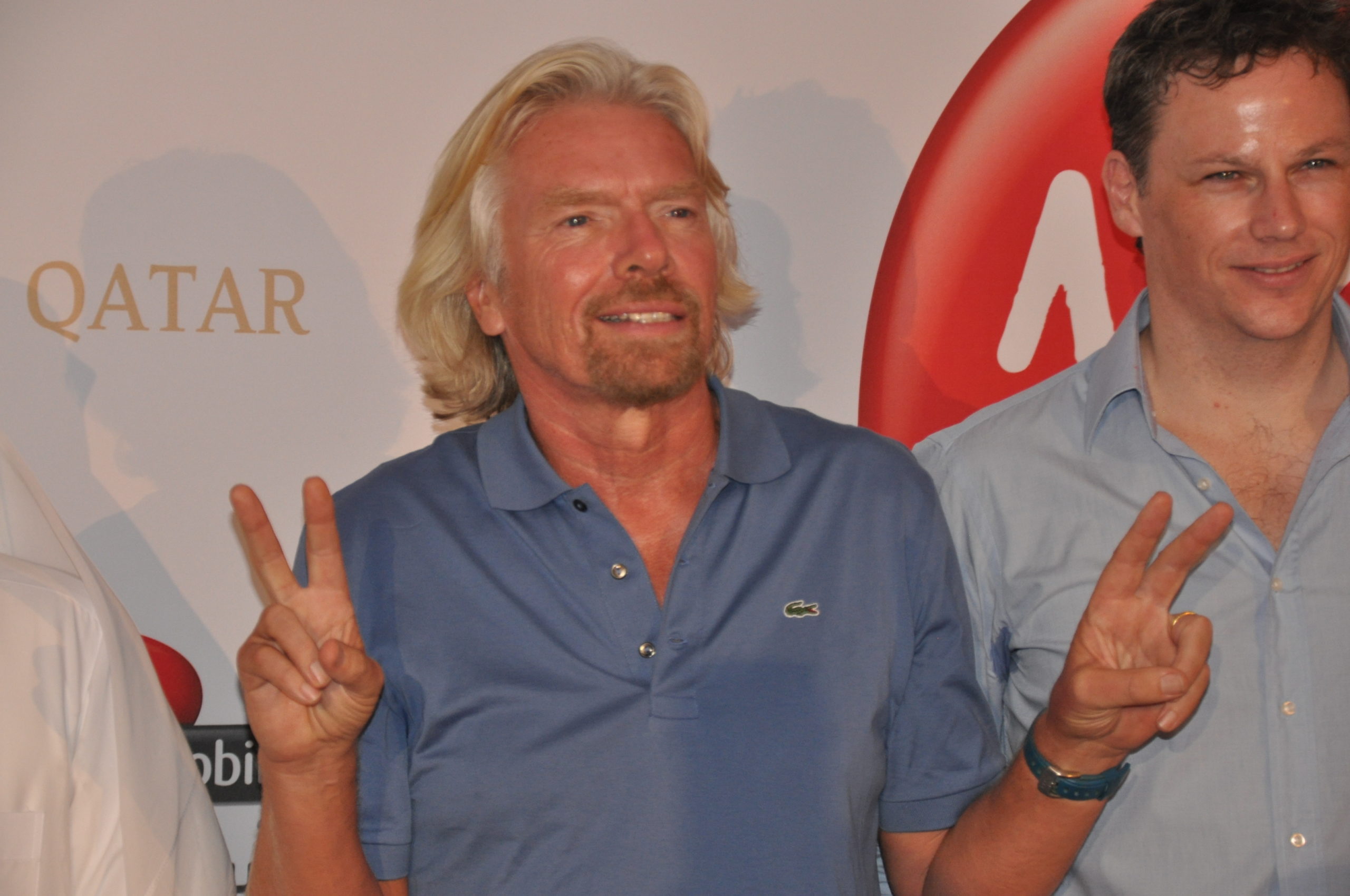 Richard Branson is standing in front of a wall with the Qatar Airways and Virgin Moblie logos on it. Two unknown people stand on either side of Branson. Branson is making V-signs with both his hands.