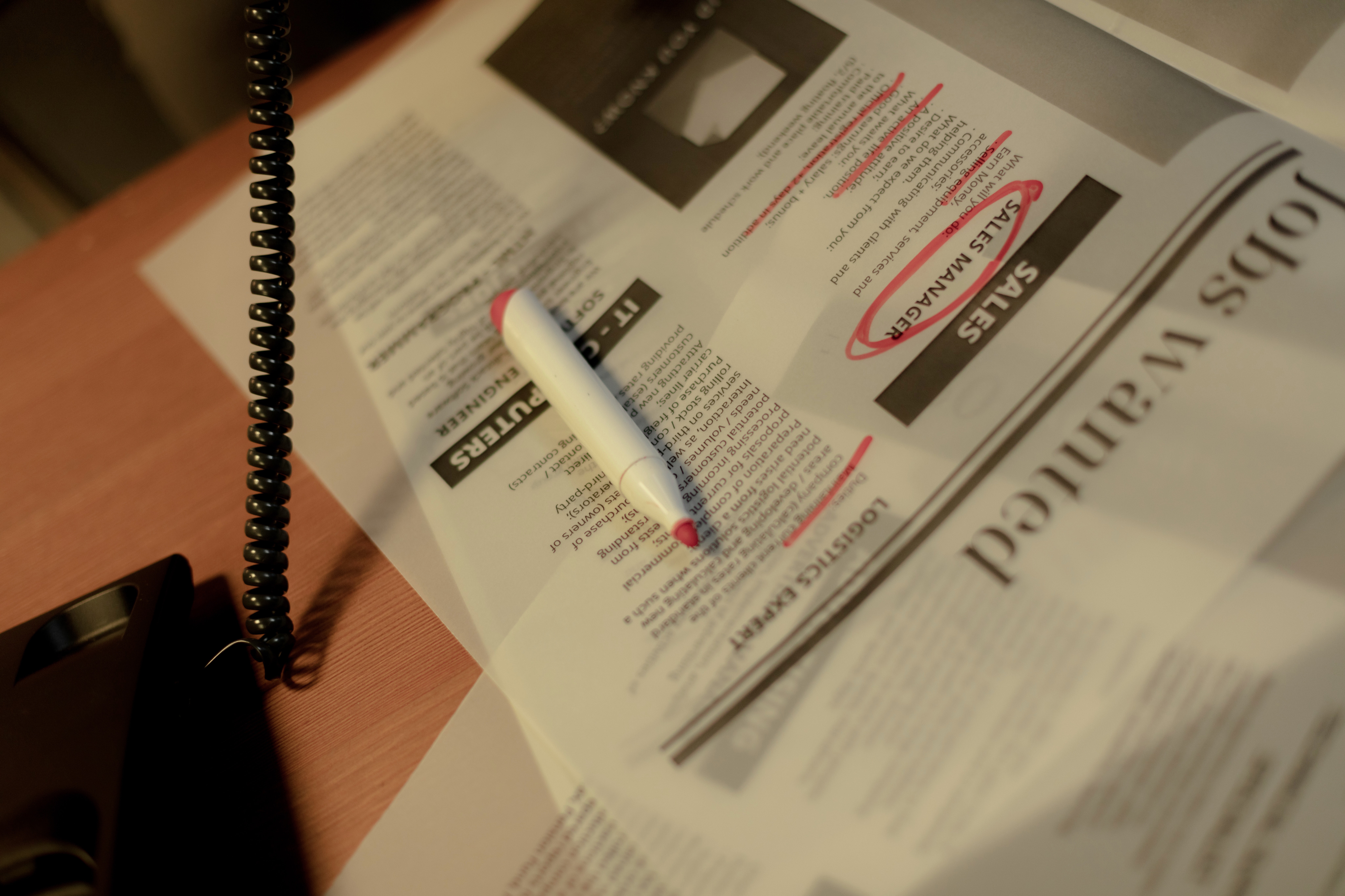 A newspaper and a red marker lie on a table. The jobs section of the newspaper is shown and the role of a sales manager is circled in red.