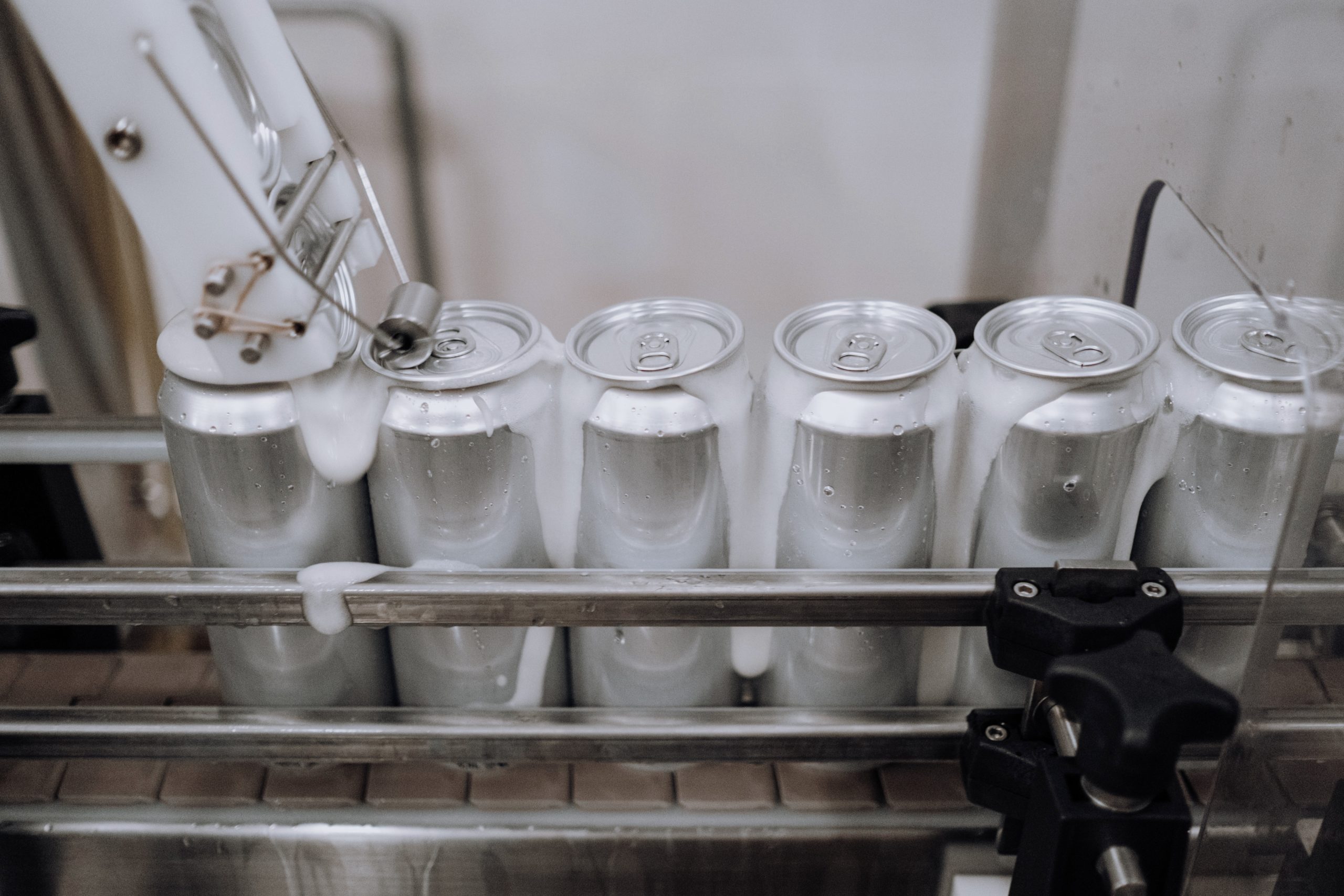 Five soft drink cans stand on a factory assembly line. Some fizzy liquid is pouring from the cans.