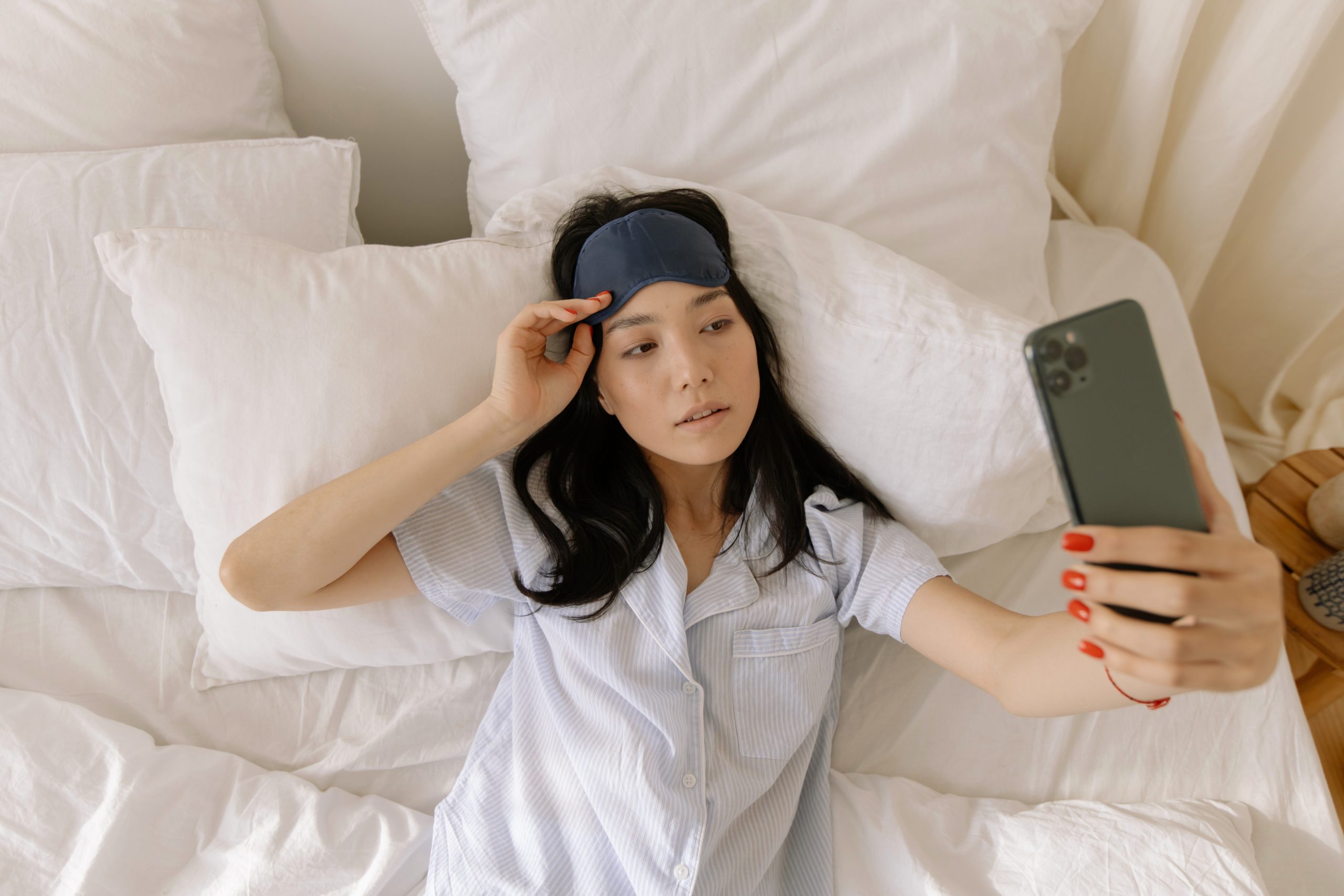 A woman is lying on her bed. She is taking a selfie on her smartphone.