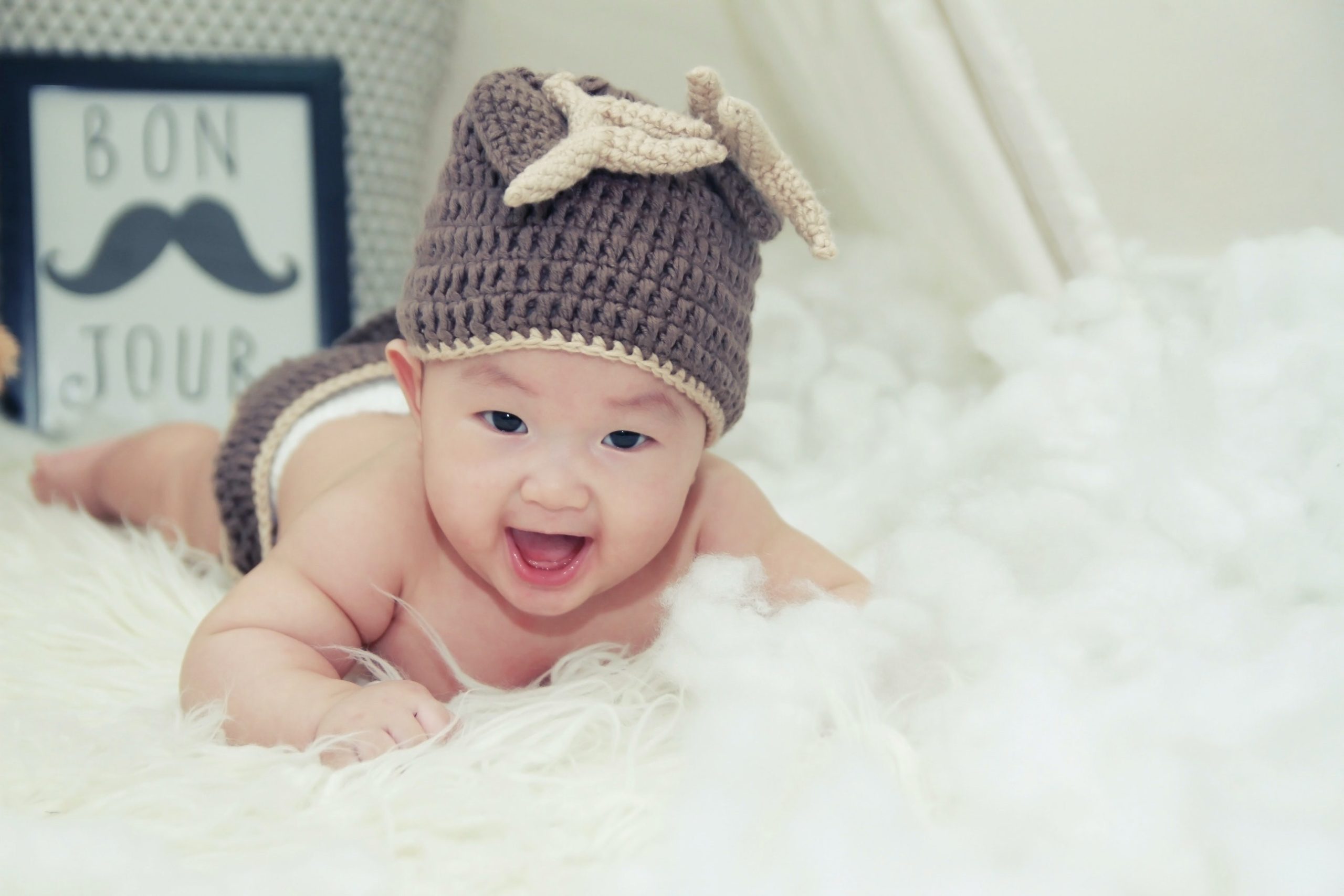A baby who is wearing a grey knitted hat is lying on a fluffy white surface. A frame with a moustache and the words, "BON JOUR,' is in the background.