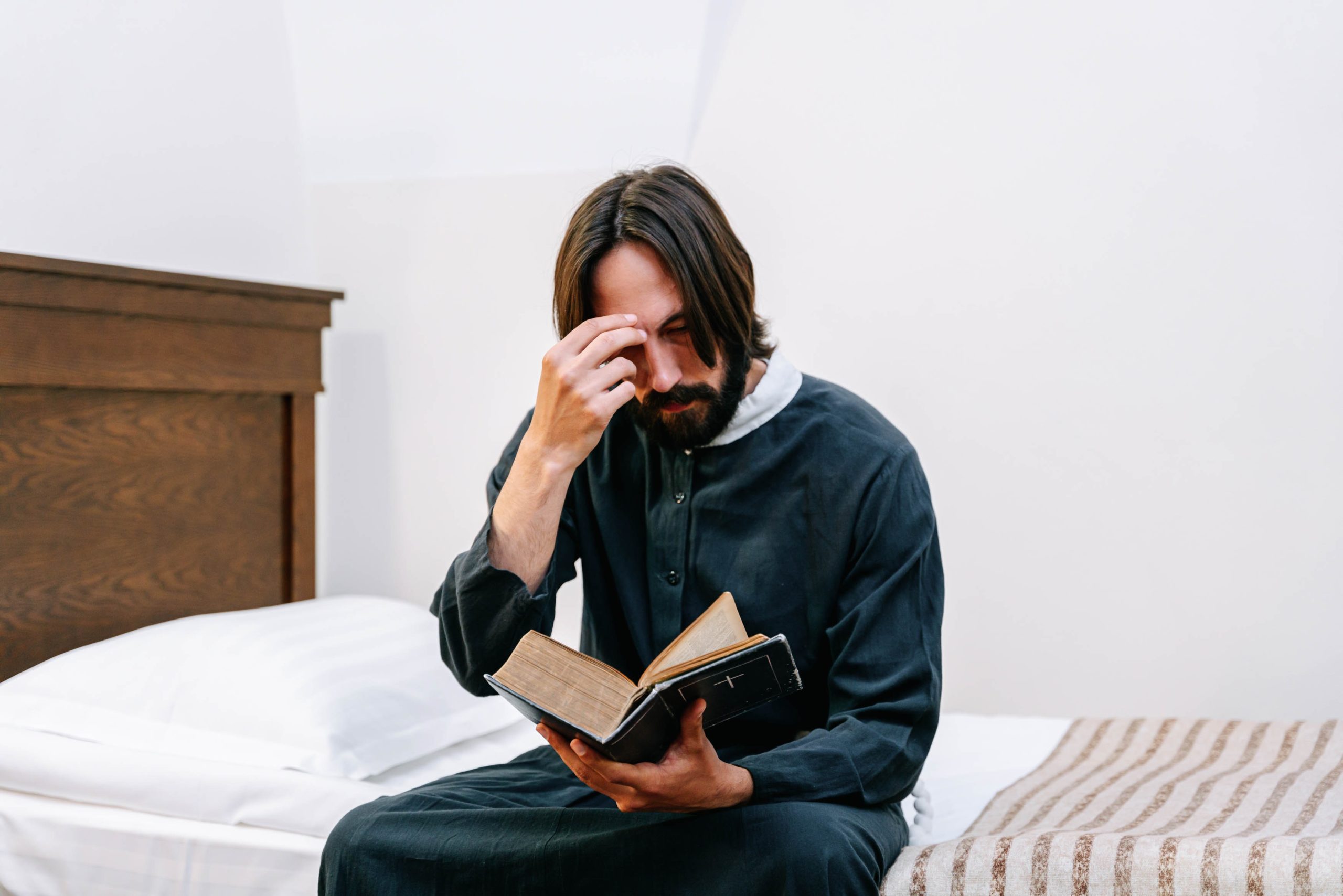 A man is sitting on a bed. He is reading a Bible and has his hand on his forehead.