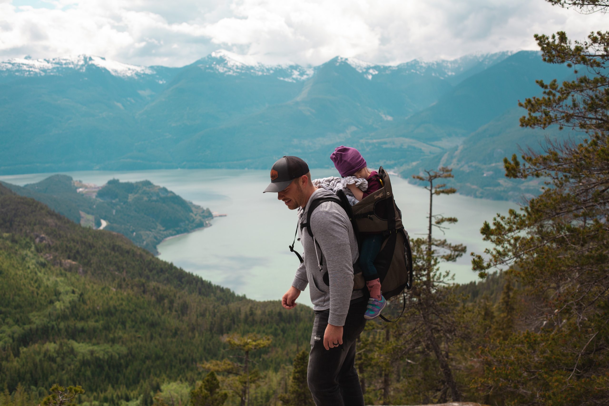 A man is carrying his child in a special rucksack and walking past a lake.