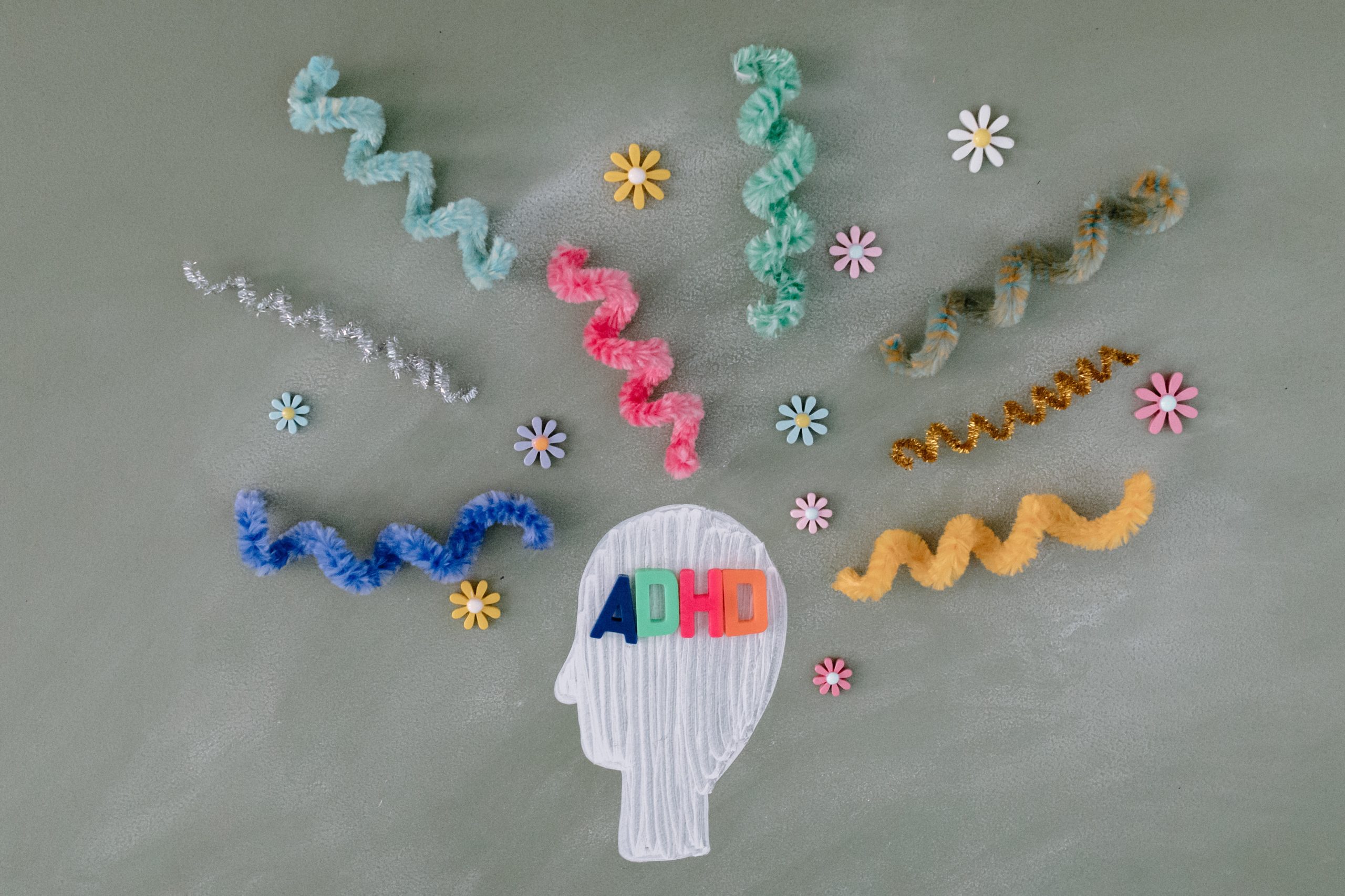 A head is drawn on a green chalkboard. Magnetic letters that spell, "ADHD,' lie on the head. Twisted pipe cleaners and small flowers surround the head.