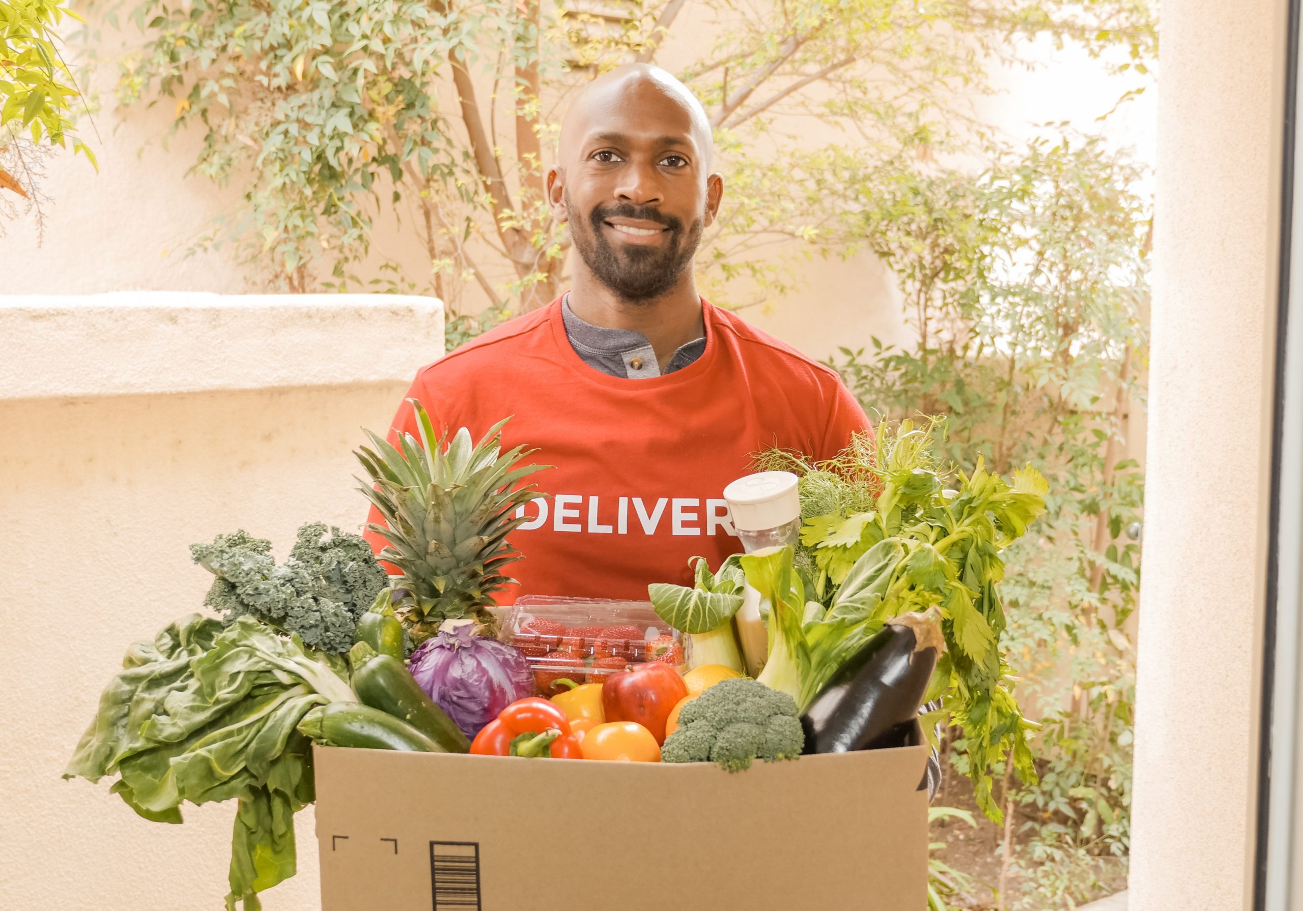A man who is wearing an orange T-shirt that says, "DELIVERY," is smiling. He is carrying a box of food.