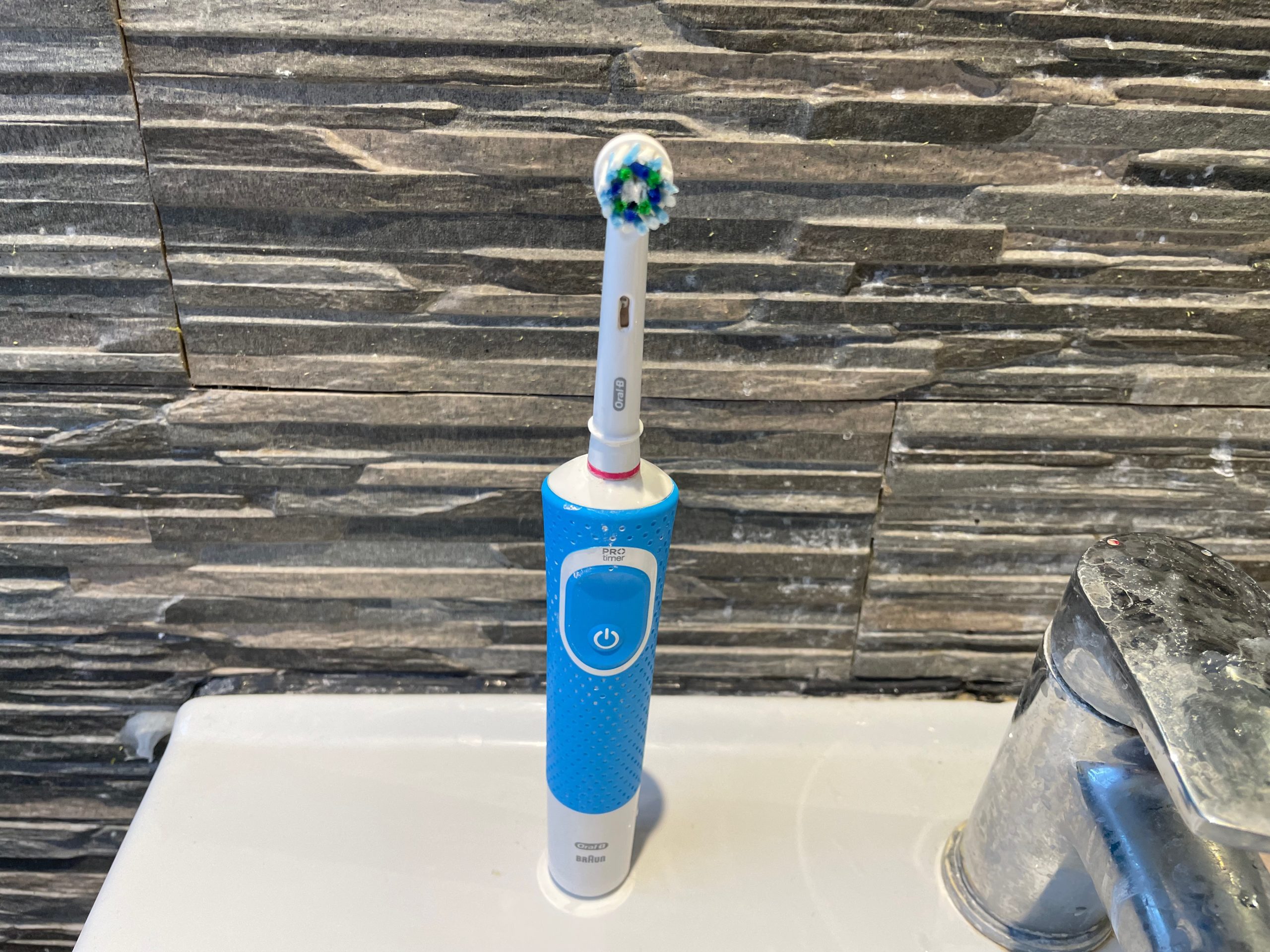 An electric toothbrush stands on a sink in front of a stone wall.