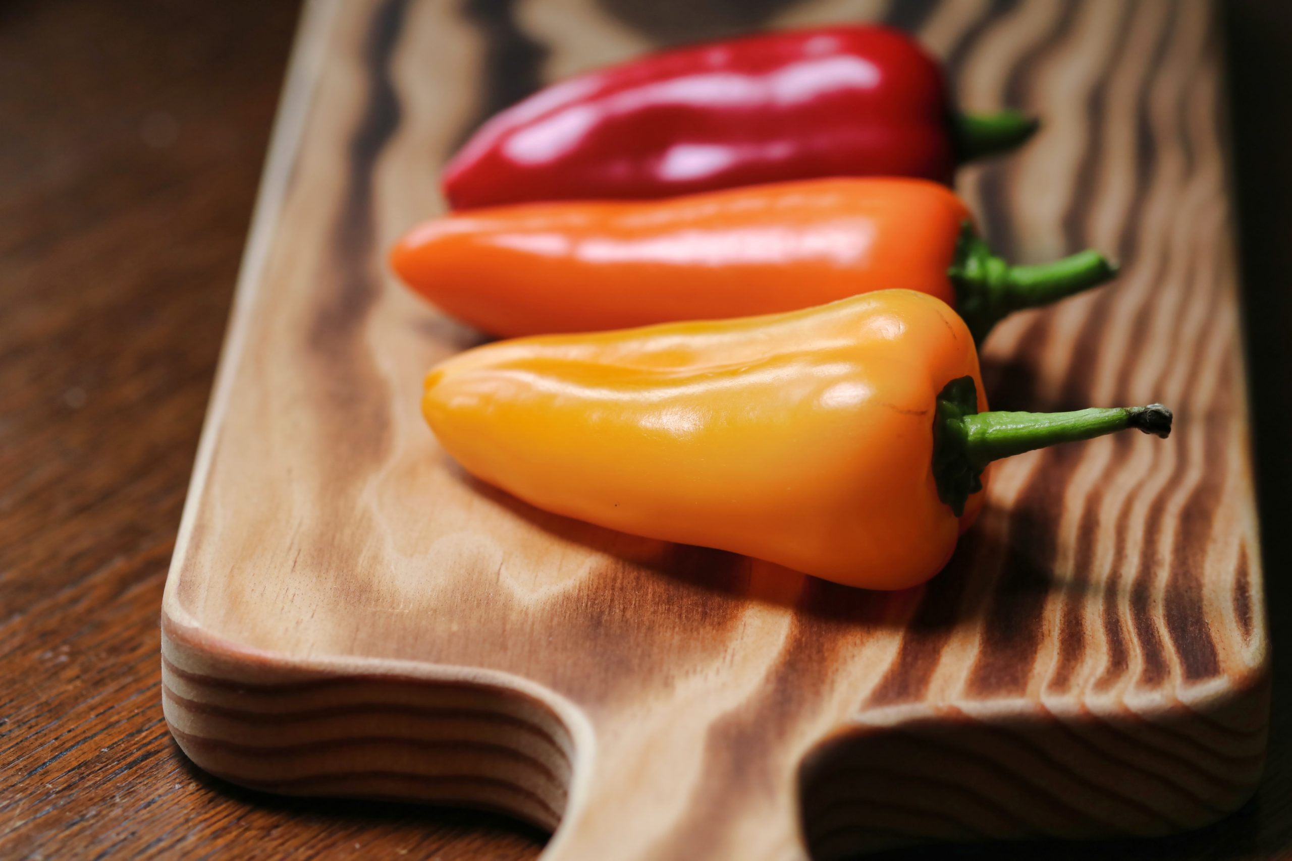 Three different coloured sweet pointed peppers lie on a wooden chopping board.
