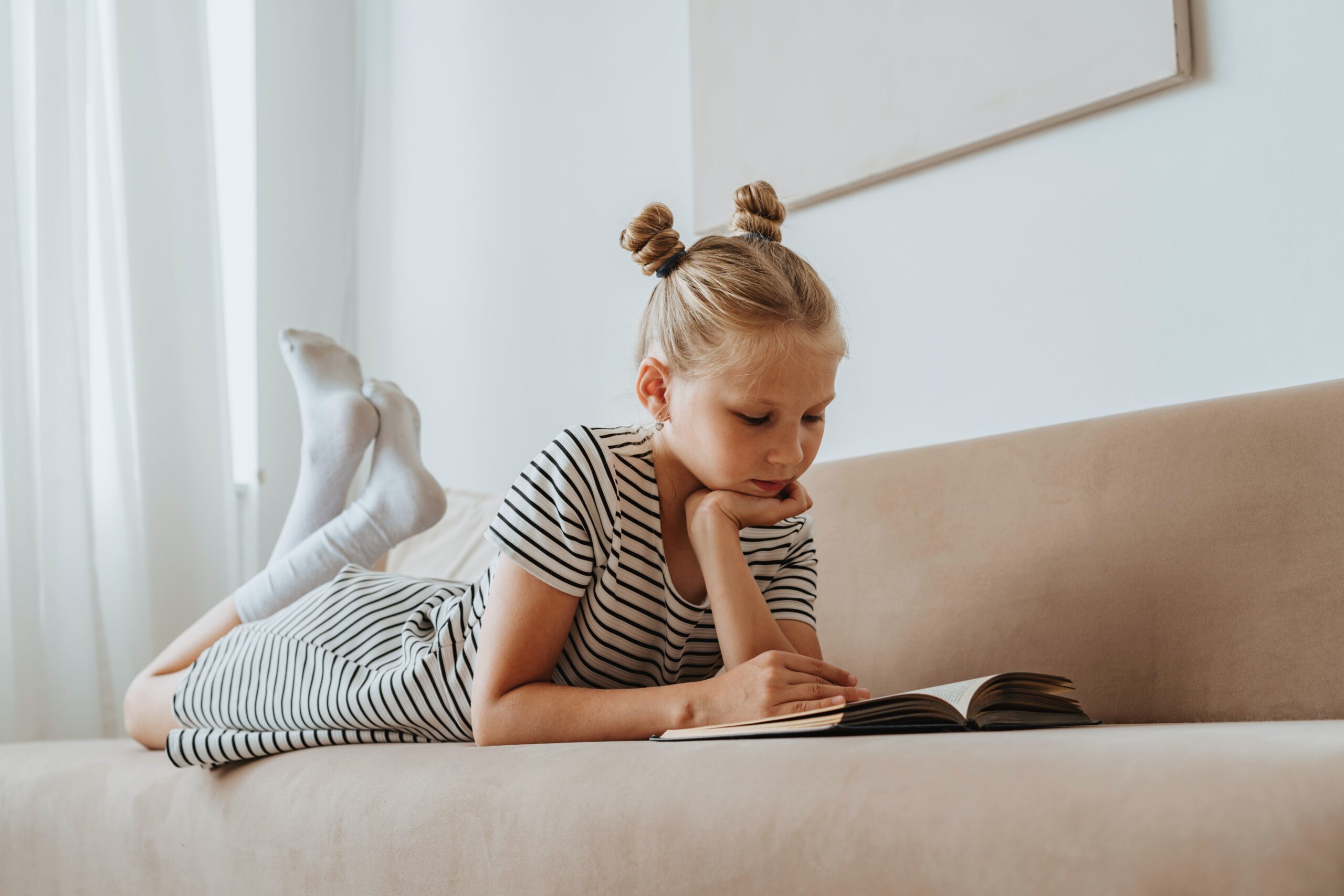 A young girl reads a book as she lies on her front on a couch.