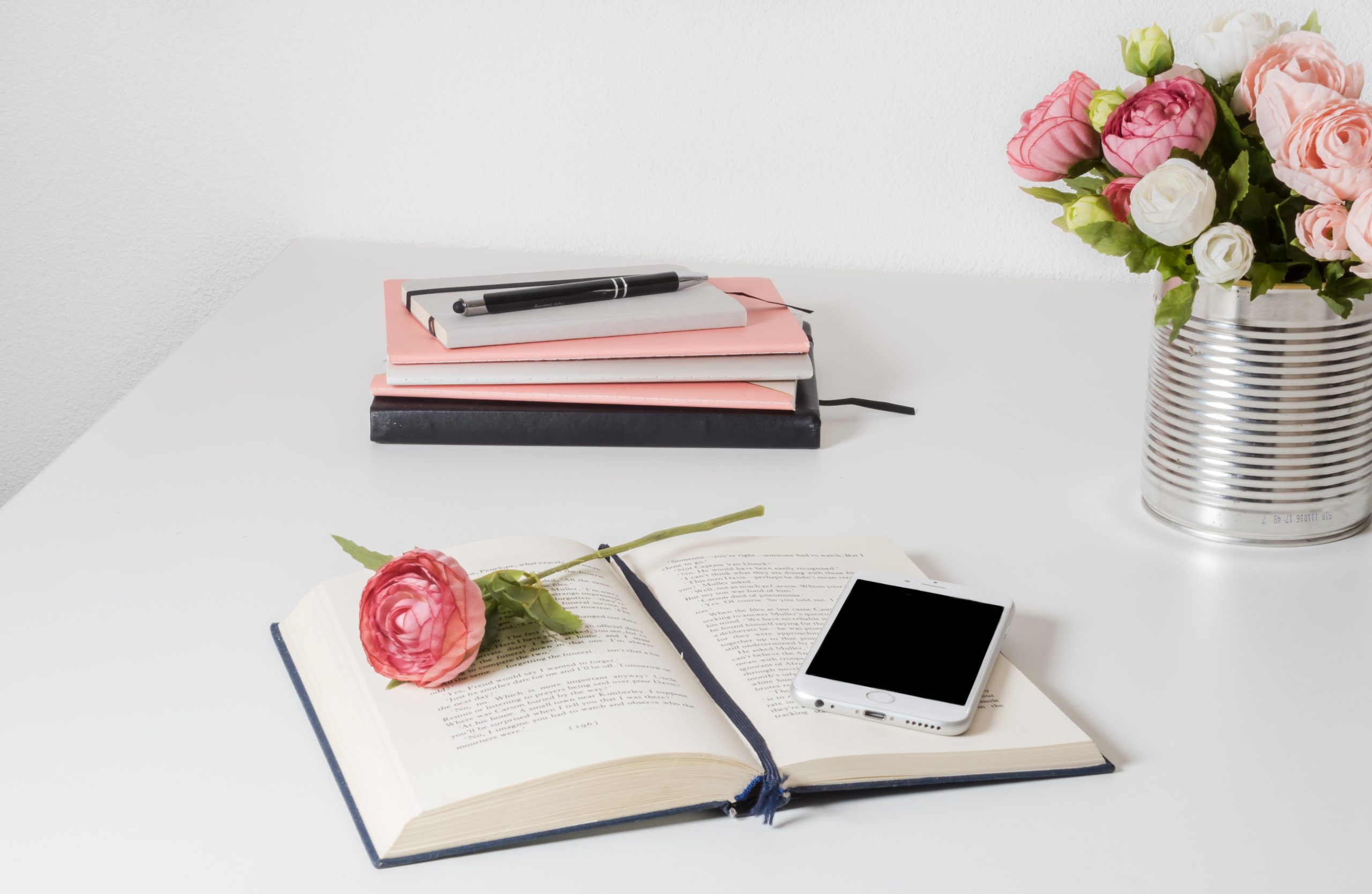 A book, a rose, a smartphone, a pile of diaries, a pen and a tin of flowers sit on a white table.