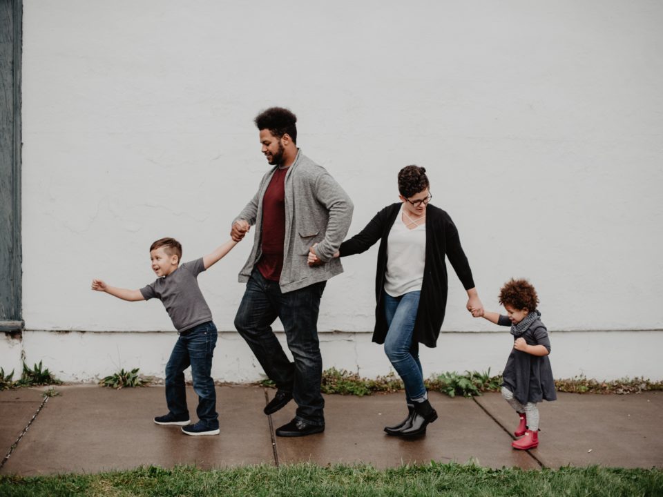 A family of four are walking down a street, holding hands and smiling.
