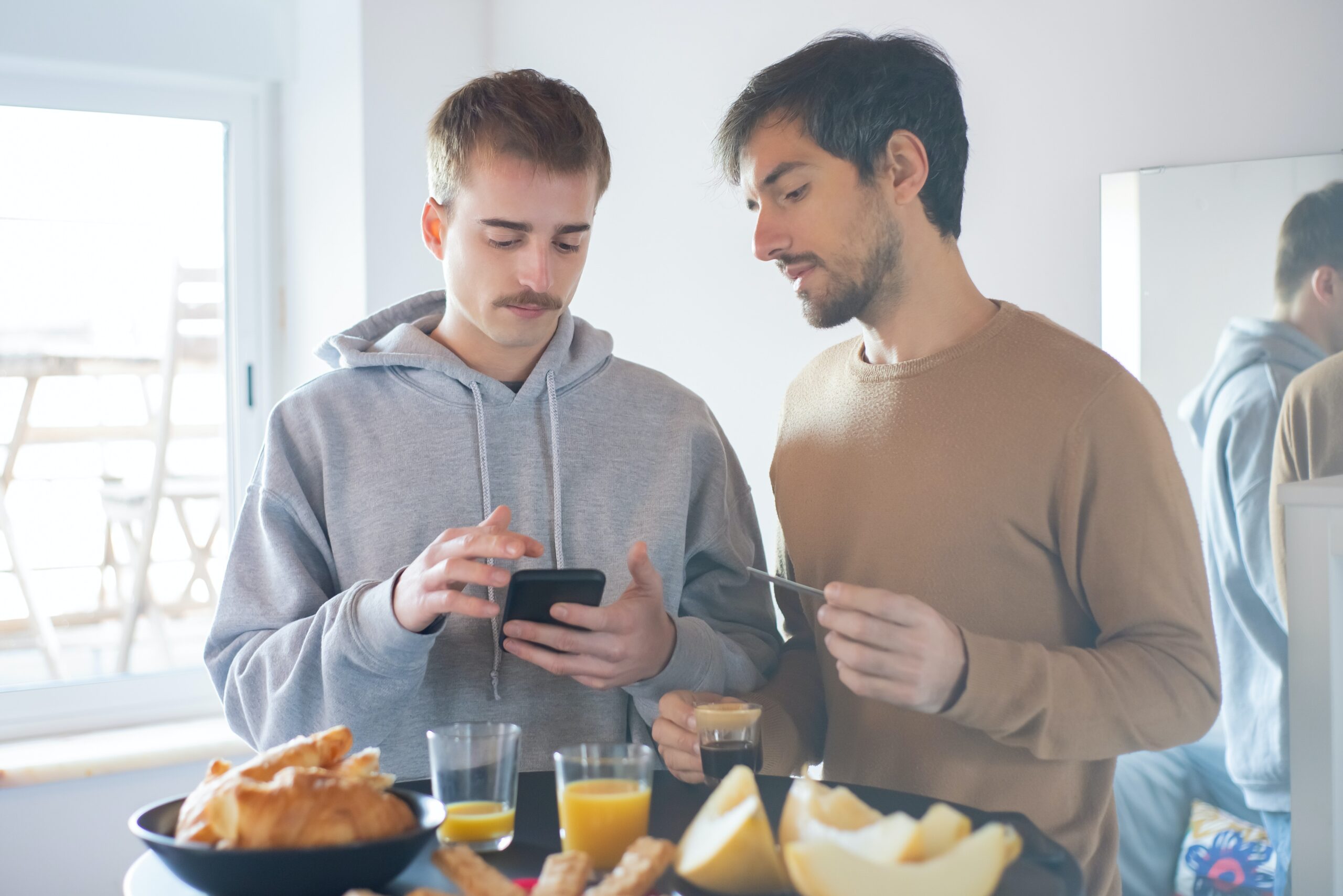Two men stand behind a table with breakfast items on it. One man uses his smartphone and the other looks at him.