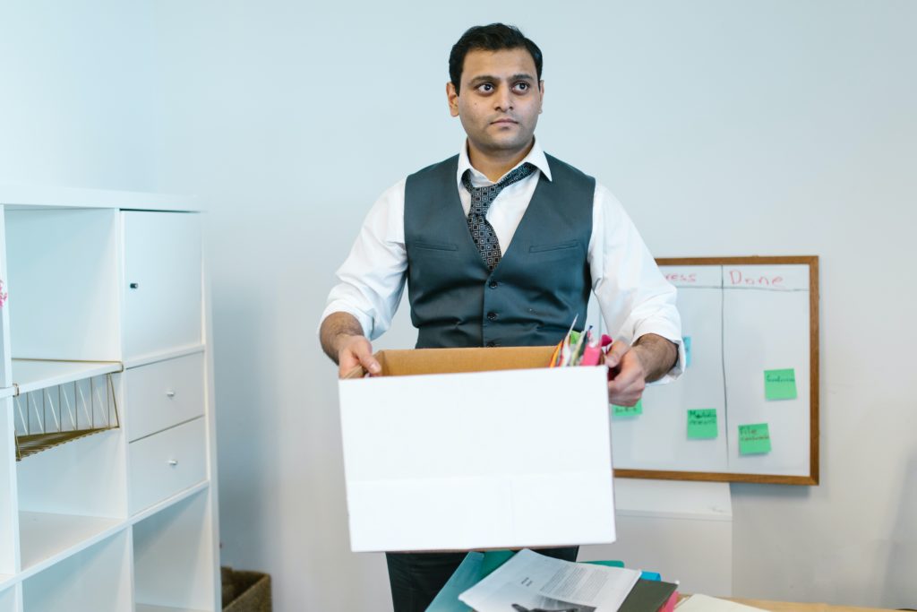 An unhappy male employee is holding a box of office supplies, which suggests that he has been fired.