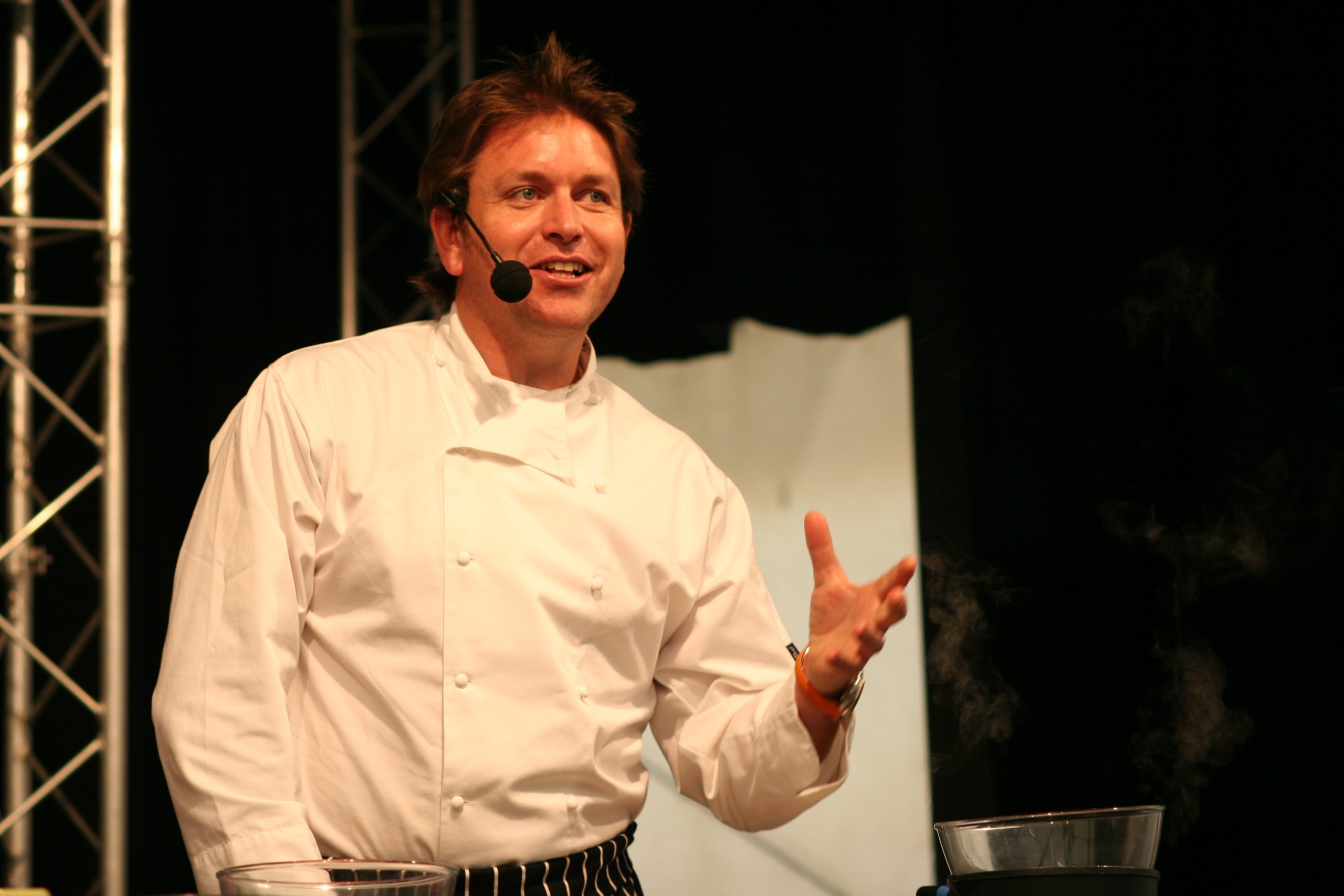 The celebrity chef James Martin is standing in front of a black and white background. He is wearing a headset and is holding up one hand.