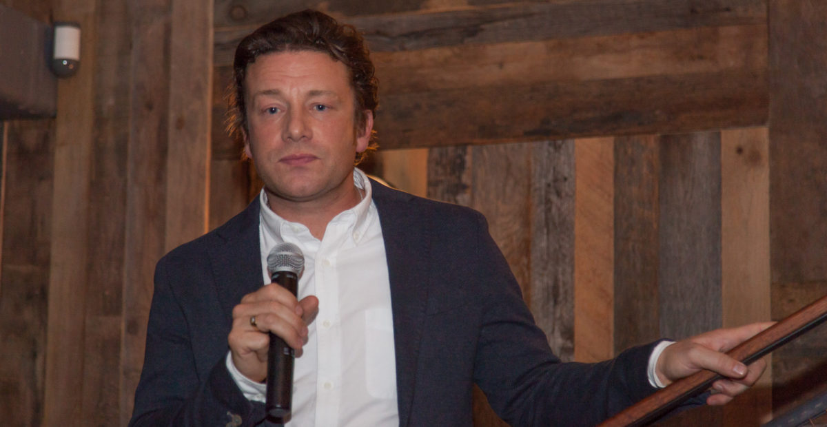 The celebrity chef Jamie Oliver is standing in front of a wooden wall. He is holding a microphone in one hand.