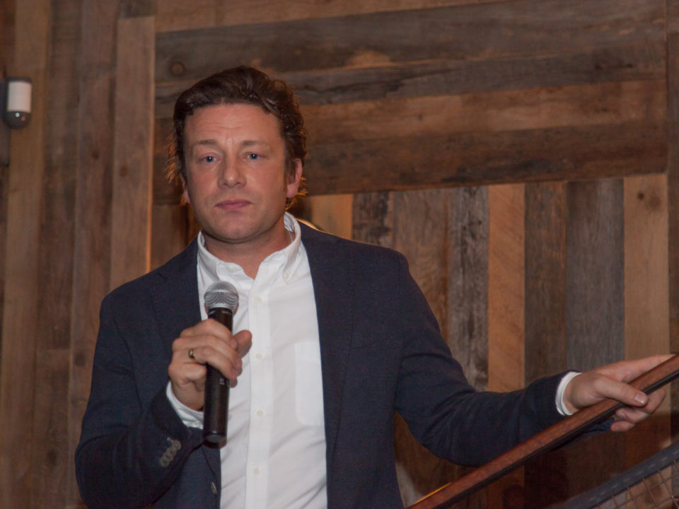 The celebrity chef Jamie Oliver is standing in front of a wooden wall. He is holding a microphone in one hand.