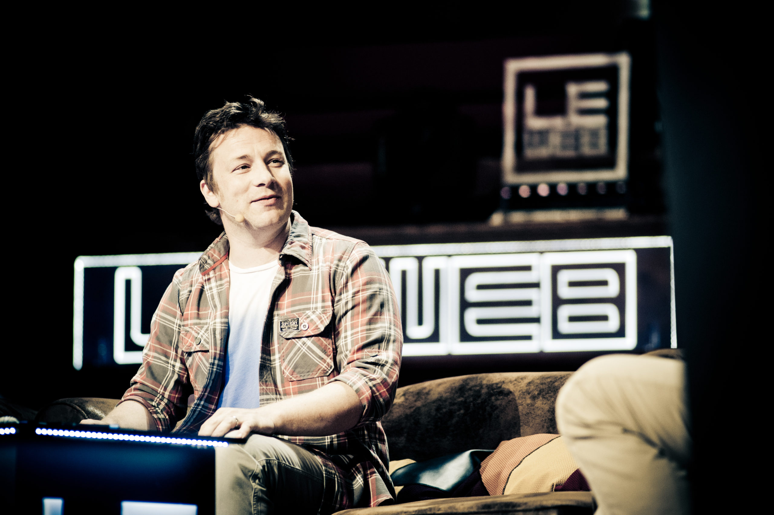 The celebrity chef Jamie Oliver is sitting on a couch. Behind him is a black background that has 2 different versions of the LEWEB logo on it. 