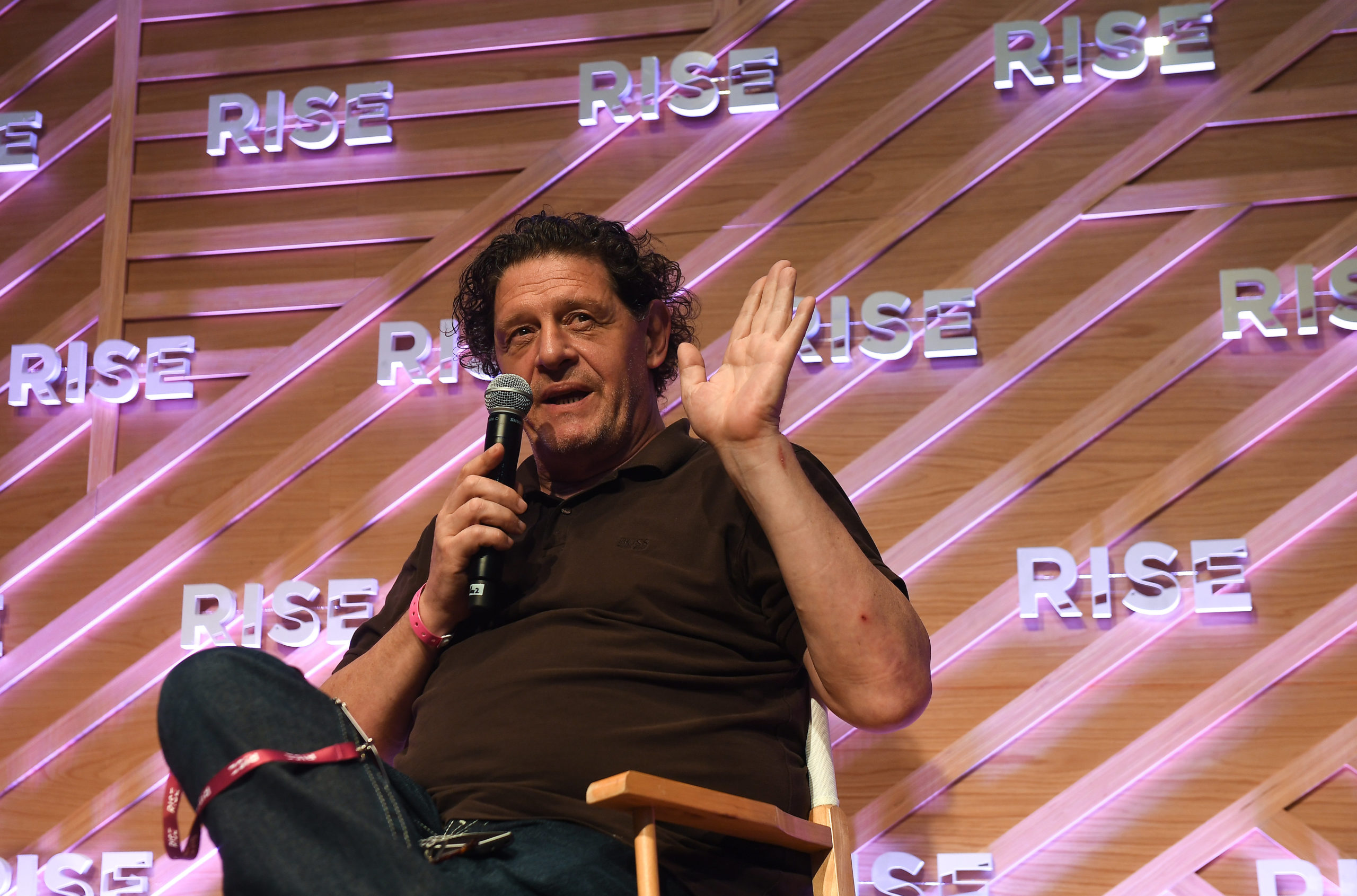 The celebrity chef Marco Pierre White is sitting in a chair. He is holding a microphone in one hand and is holding up the other palm. Behind him is a diagonal striped pink and brown wall with the RISE logo all over it.