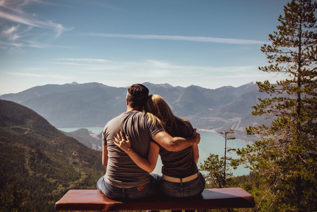 A backshot of a man and a woman facing some trees, some mountains and a lake. The man and woman have their arms across each other's backs. 