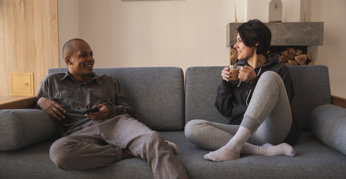 A couple sit on a couch and smile at each other. The man is holding a smartphone and the woman is holding a mug.