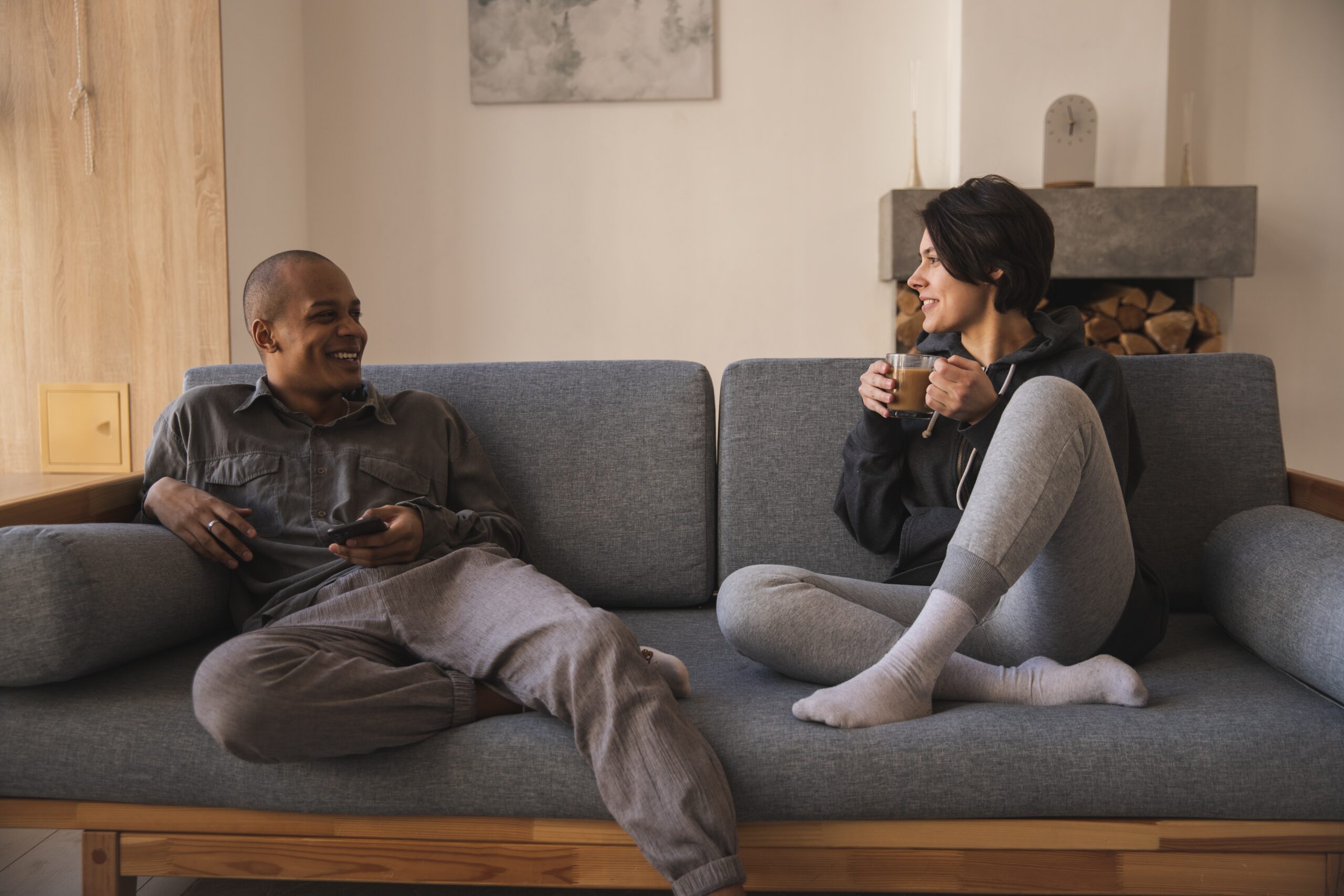A couple sit on a couch and smile at each other. The man is holding a smartphone and the woman is holding a mug.